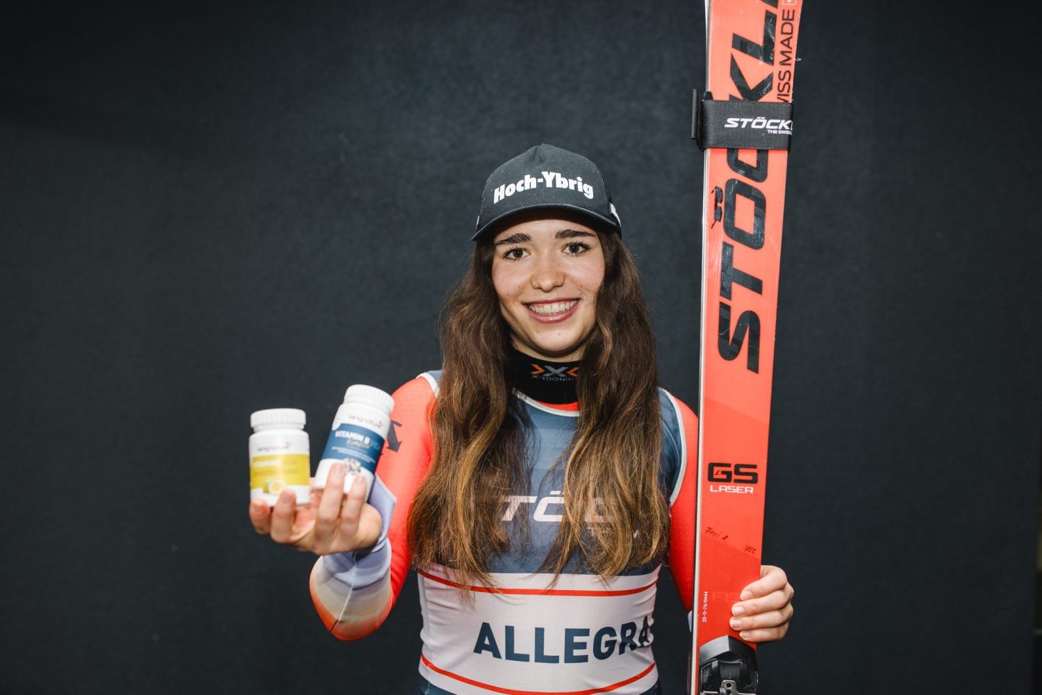 Eine lächelnde Tennisspielerin posiert mit einem Schläger auf der Schulter und wirft eine kleine Flasche mit der Aufschrift The Zinc by kingnature, eine Nahrungsergänzung aus der Schweiz, vor einen schwarzen Hintergrund.