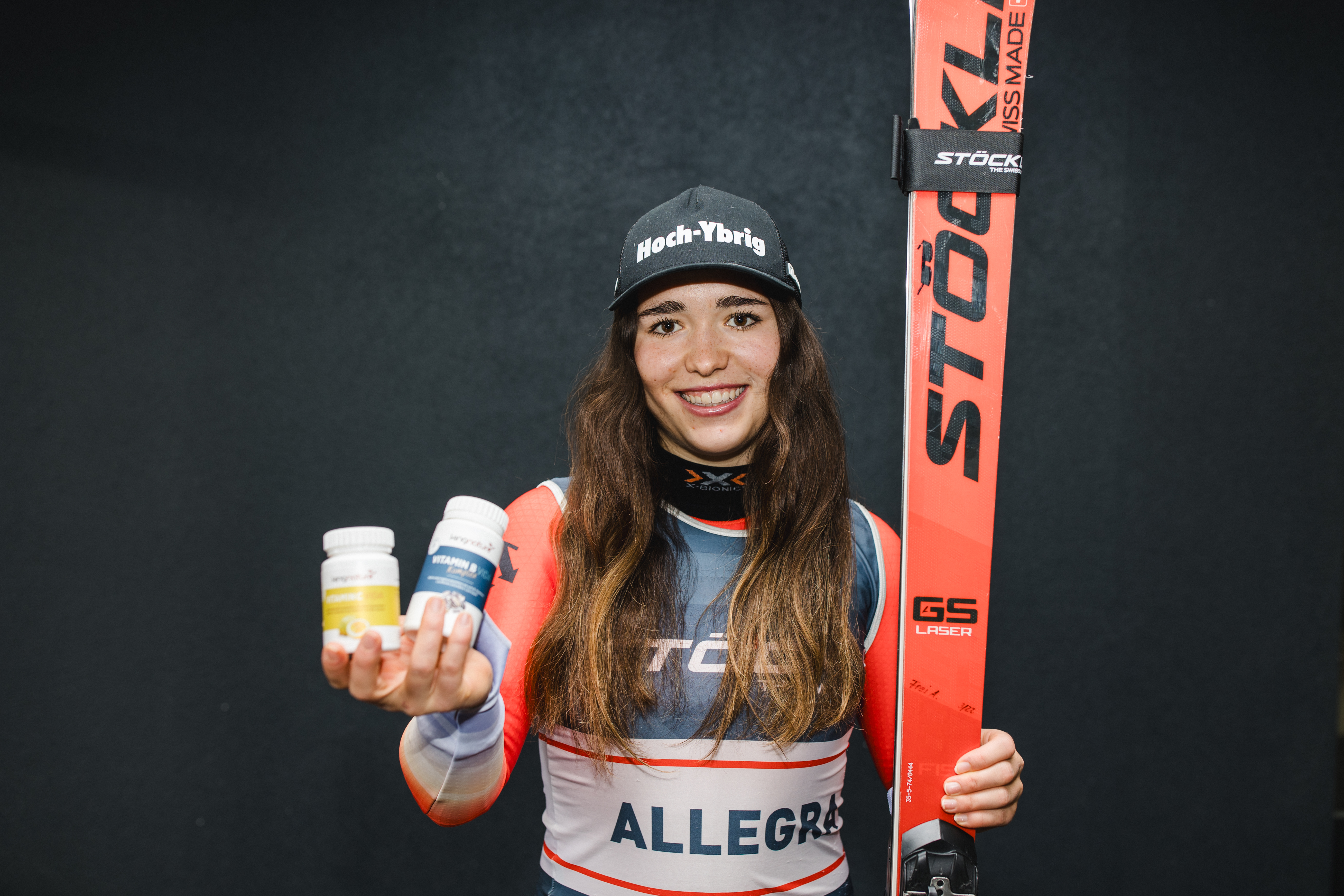Vor einem schlichten, dunklen Hintergrund steht eine lächelnde Allegra Frei in Rennkleidung, die in der einen Hand drei Getränkeflaschen und in der anderen einen roten Stockli-Ski hält.
