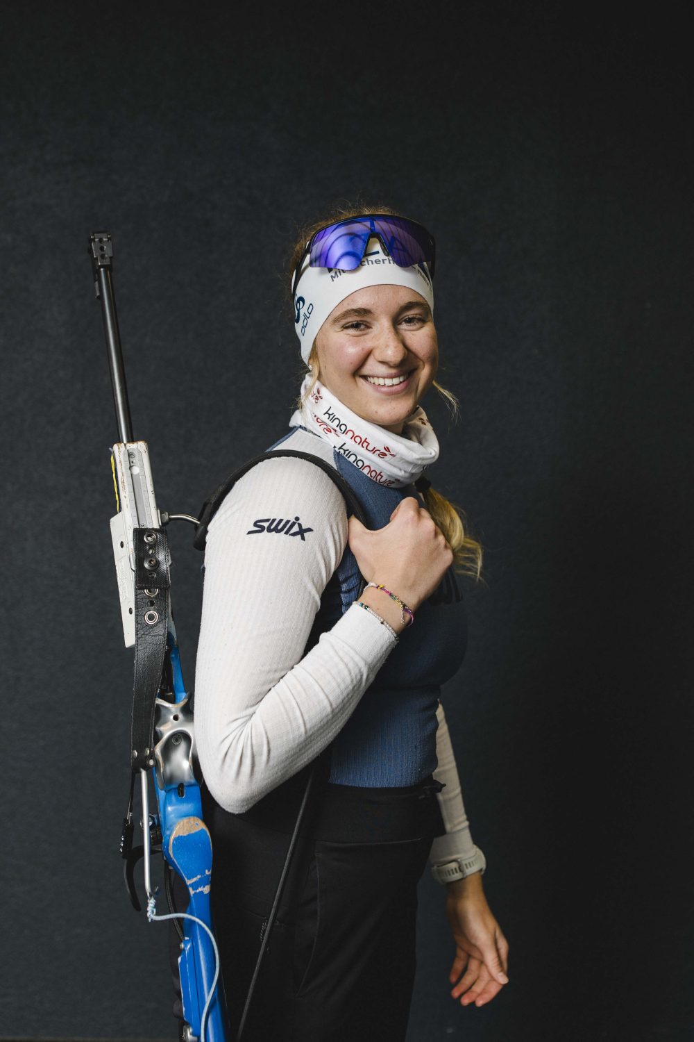 Eine lächelnde Frau in einem schwarz-grünen Sportdress hält einen Unihockeyschläger und zeigt vor einem schwarzen Hintergrund eine Flasche mit Amino Vida-Ergänzungsmitteln.