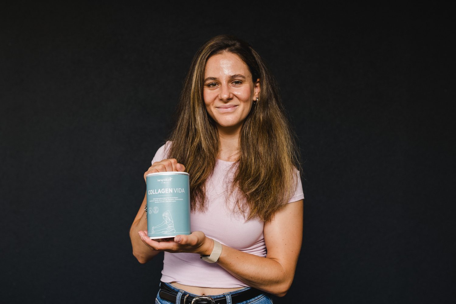 Talina Gantenbein, mit langen braunen Haaren, trägt ein hellrosa T-Shirt und Jeans, während sie vor einem dunklen Hintergrund steht, lächelt und einen Behälter mit der Aufschrift „Collagen Vida“ in der Hand hält.