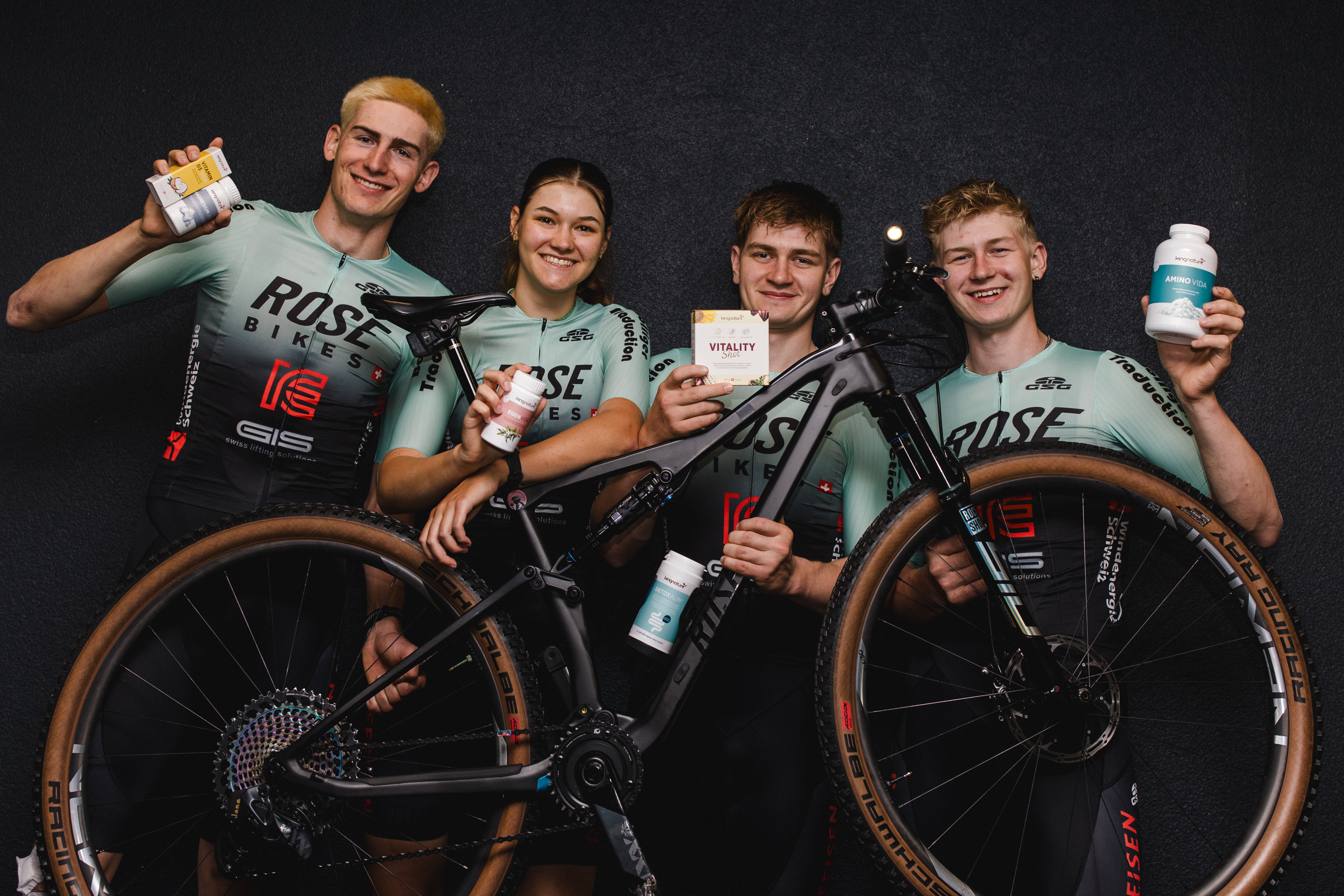 Vier Radfahrer des ROSE Bikes Factory Racing Teams stehen vor einem schwarzen Fahrrad, lächeln und halten vor einem dunklen Hintergrund verschiedene Flaschen und Päckchen mit Nahrungsergänzungsmitteln in die Kamera. Als stolze Vertreter der Schweiz verkörpern sie Zusammenhalt und Teamgeist.