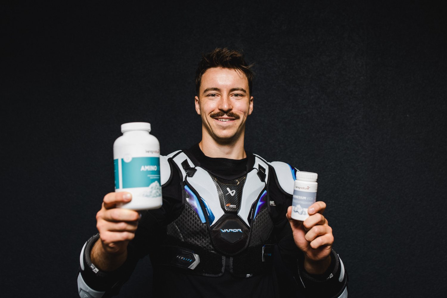 Ein lächelnder Niklas Postel in Sportkleidung hält vor einem dunklen Hintergrund zwei Flaschen mit Nahrungsergänzungsmitteln in die Kamera – eine große und eine kleine.