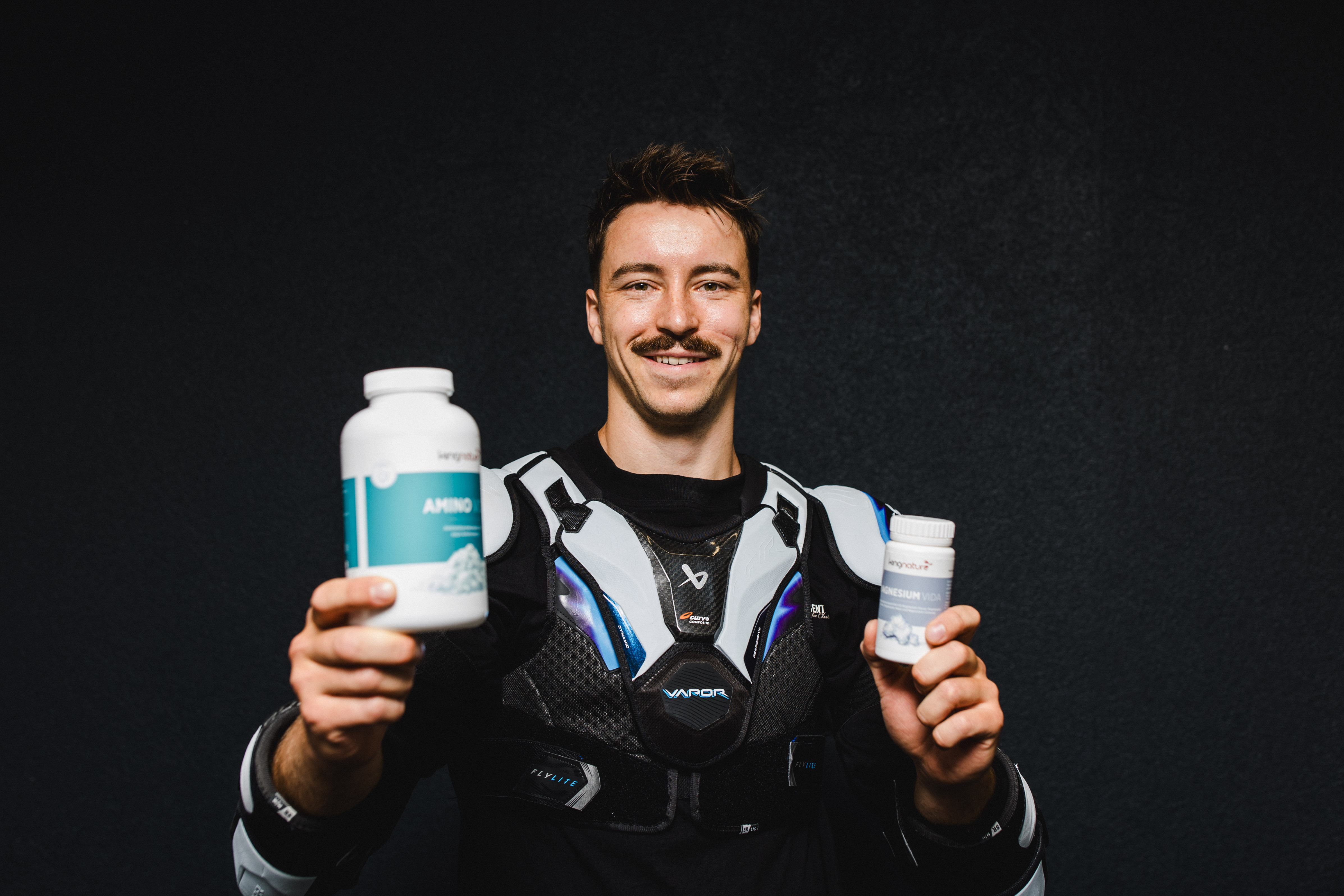 Ein lächelnder Niklas Postel in Sportkleidung hält vor einem dunklen Hintergrund zwei Flaschen mit Nahrungsergänzungsmitteln in die Kamera – eine große und eine kleine.