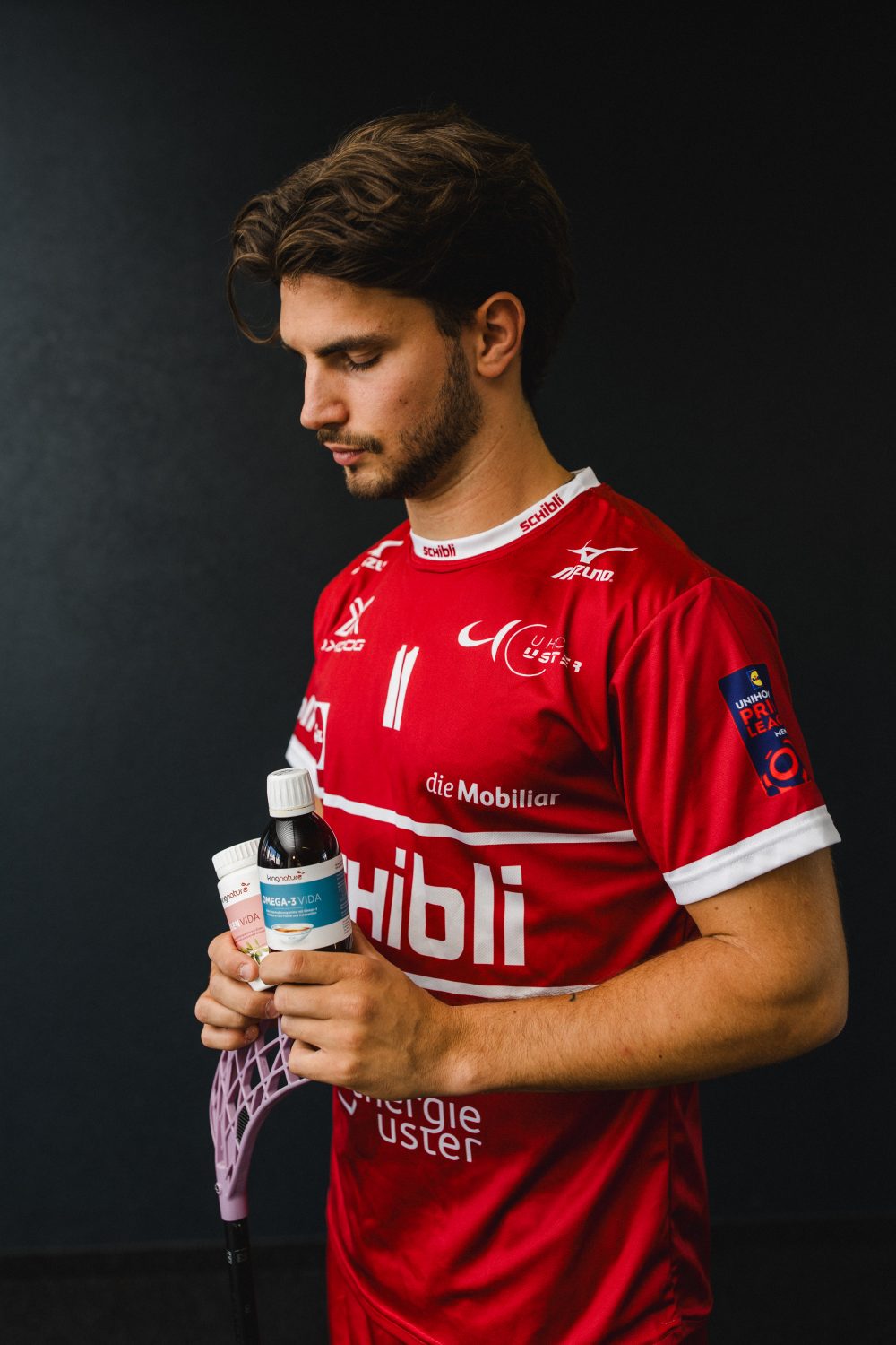 Ein junger Mann in einem roten Sporttrikot hält einen Floorballschläger und eine Flasche mit einem Sportgetränk in der Hand und steht vor einem dunklen Hintergrund – das Bild fängt die entschlossene Ausstrahlung von Josia Pfister ein.