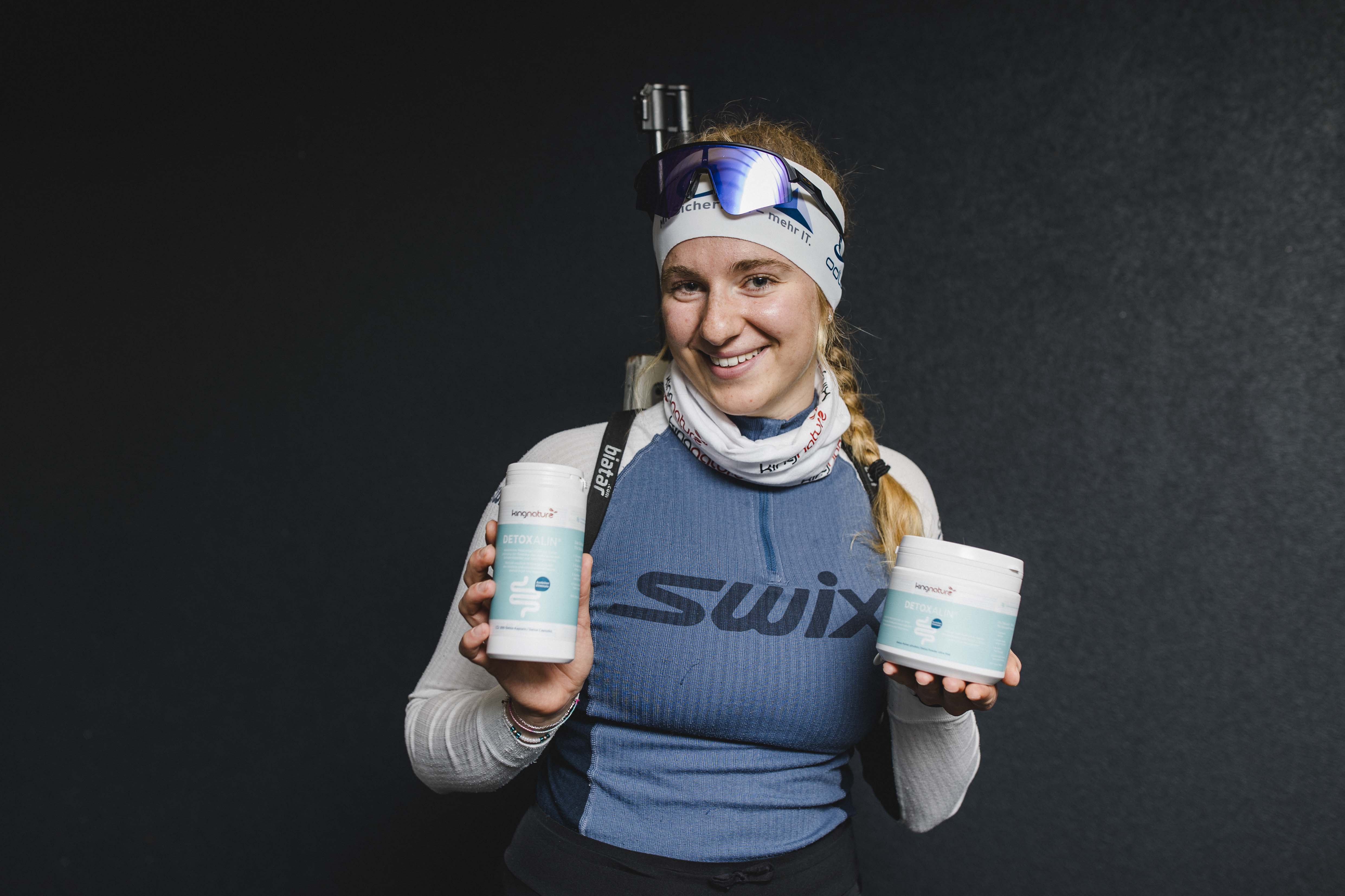 Lisa Naegeli, eine lächelnde Frau in Sportkleidung und mit Stirnband, hält vor einem dunklen Hintergrund zwei Behälter mit Nahrungsergänzungsmitteln. Sie hat lange blonde Haare zu einem Zopf geflochten und trägt eine Skibrille.
