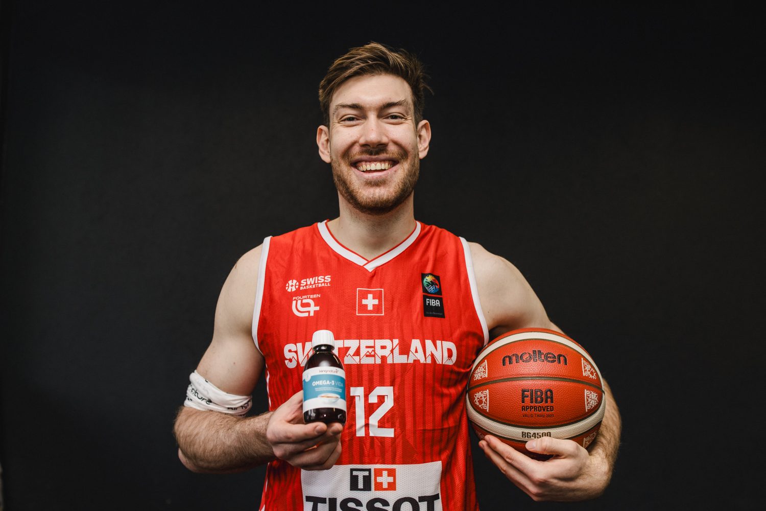 Ein lächelnder Basketballspieler, Laurent Zoccoletti, in einem roten Schweiz-Trikot, hält einen Basketball in der einen und eine Flasche mit Nahrungsergänzungsmitteln in der anderen Hand und steht vor einem schlichten schwarzen Hintergrund.