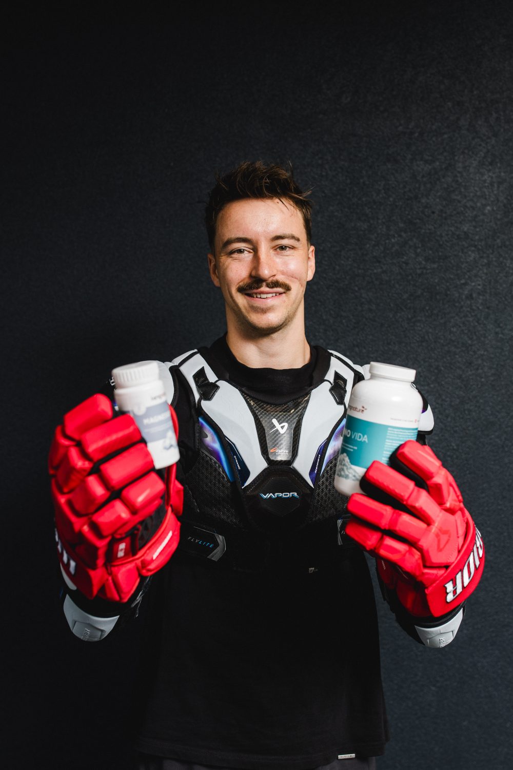 Niklas Postel, der rote Eishockeyhandschuhe und einen Brustschutz trägt, hält zwei Flaschen mit Sportgetränk in der Hand und lächelt vor einem dunklen Hintergrund in die Kamera.