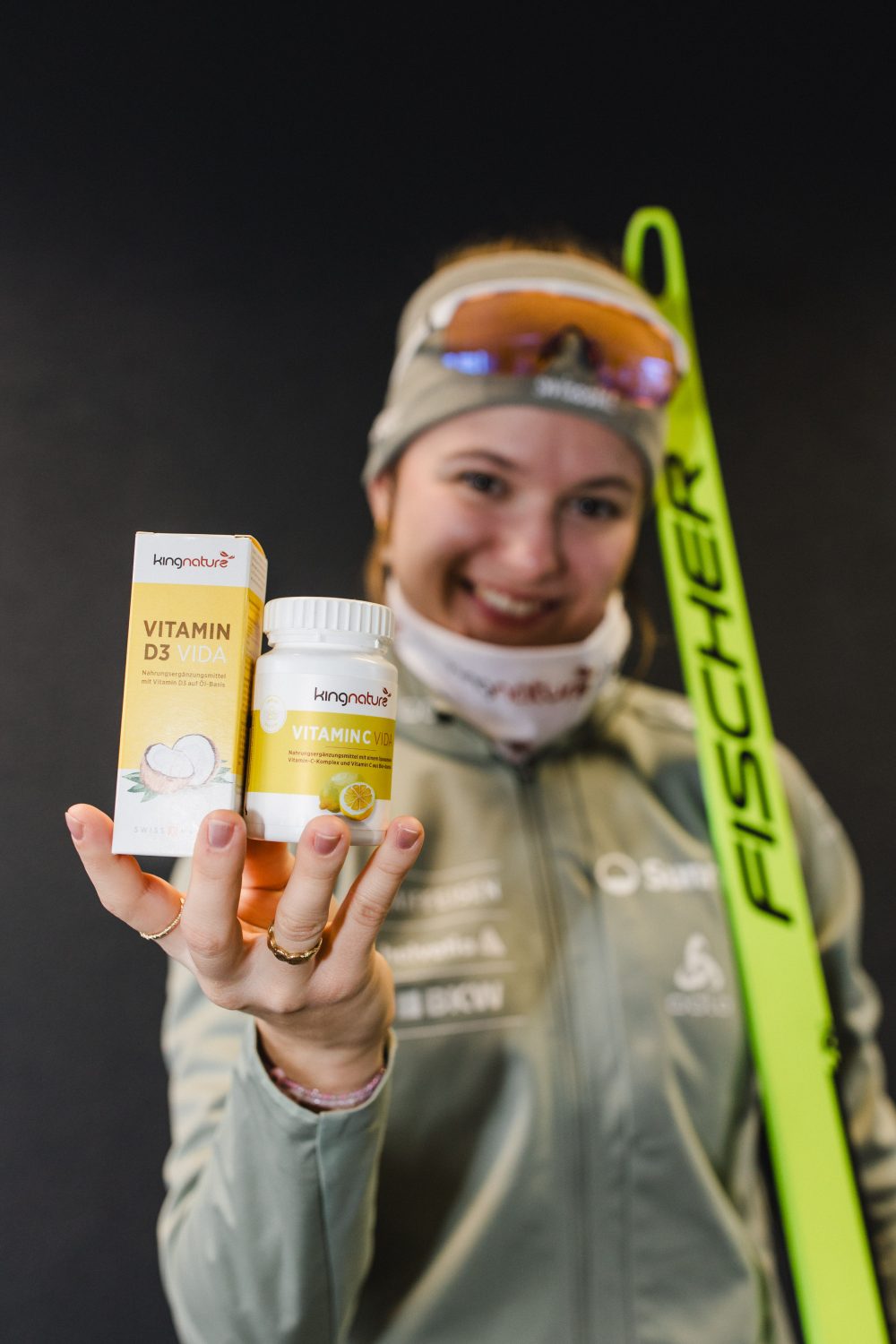 Ein lächelnder Sportler in Wintersportkleidung hält eine Flasche und eine Schachtel mit den Nahrungsergänzungsmitteln Kangavital Vitamin D3 Vida und Vitamin C Vida mit einem gelben Fischer-Ski vor einem dunklen Hintergrund in die Höhe.