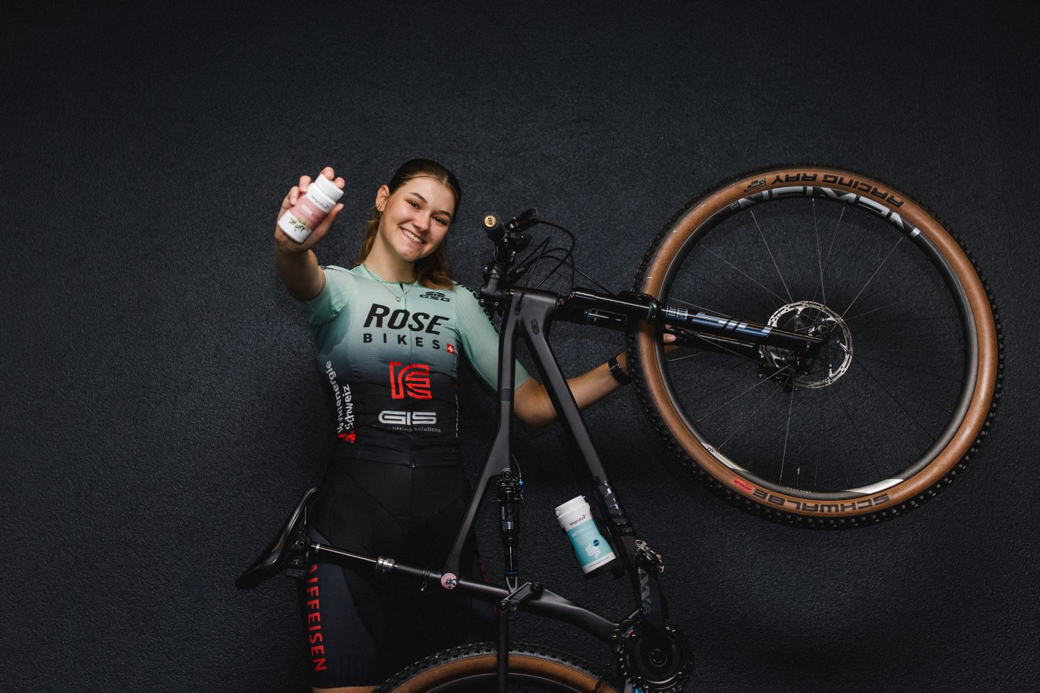 Ein Radfahrer in Rennbekleidung lächelt, hält eine Trinkflasche in der Hand und hebt ein schwarzes ROSE-Bike mit hellbraunen Reifen an. Eine weitere Trinkflasche ist am Rahmen befestigt. Das Logo des in der Schweiz ansässigen Factory Racing Teams hebt sich deutlich vom schlichten dunklen Wandhintergrund ab.