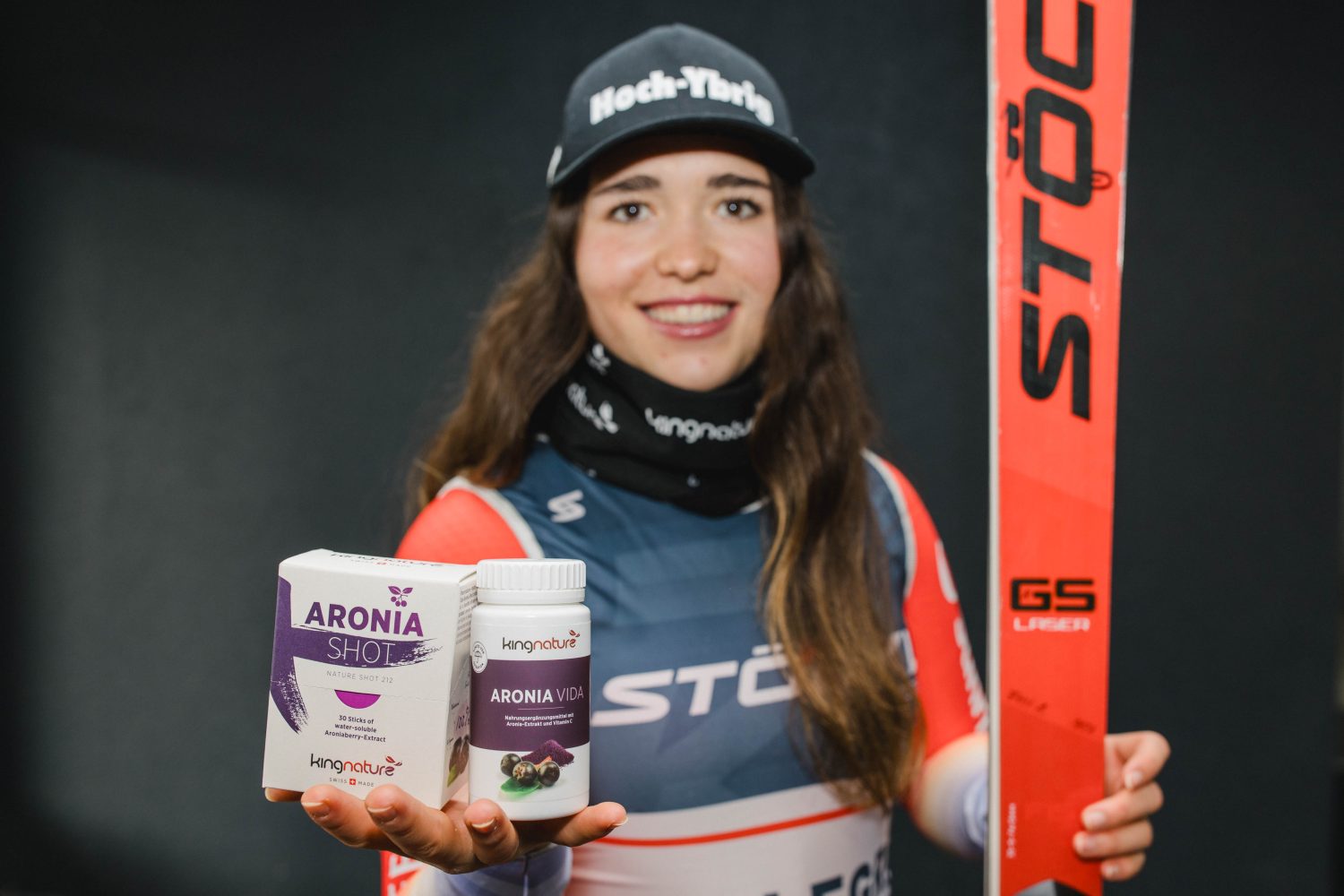Ein lächelnder Sportler in Skikleidung und schwarzer Mütze hält vor dunklem Hintergrund in der einen Hand zwei Aronia Vida Produkte - ideal für alle, die Aronia Vida kaufen wollen - und in der anderen einen roten STÖCKLI Ski.