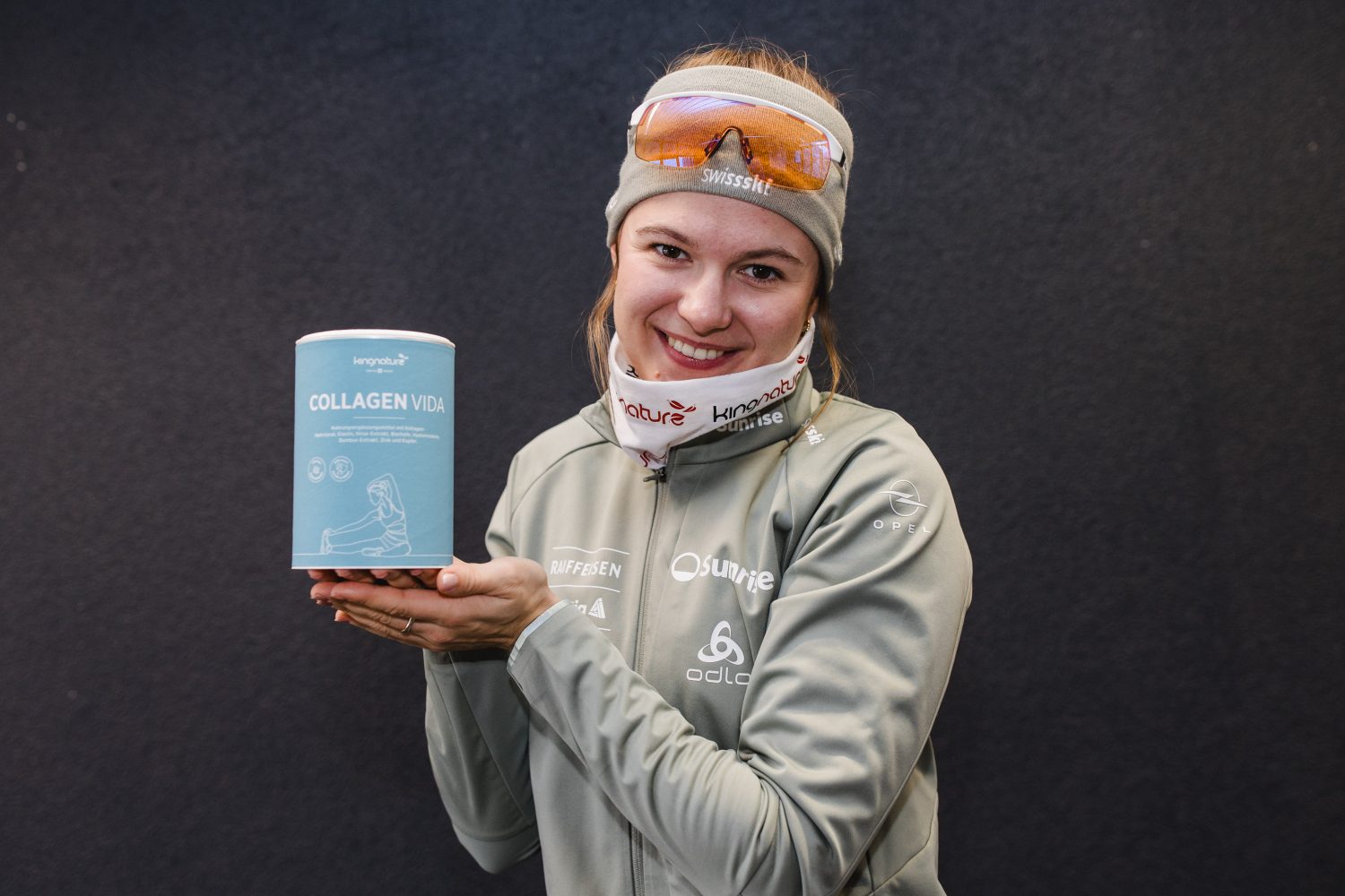 Eine lächelnde Frau in Sportkleidung und mit Stirnband hält einen Behälter mit Collagen Vida vor einem dunklen Hintergrund.