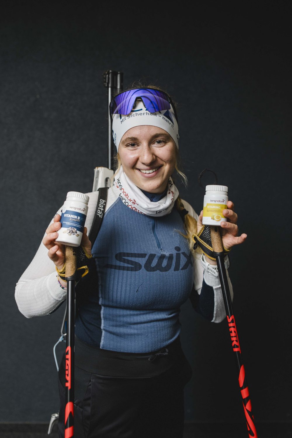Lisa Naegeli, eine Langläuferin, lächelt in Sportkleidung und mit Stirnband in die Kamera. Sie hält zwei Flaschen mit Nahrungsergänzungsmitteln in der Hand, trägt außerdem Skistöcke und ein Gewehr auf dem Rücken.