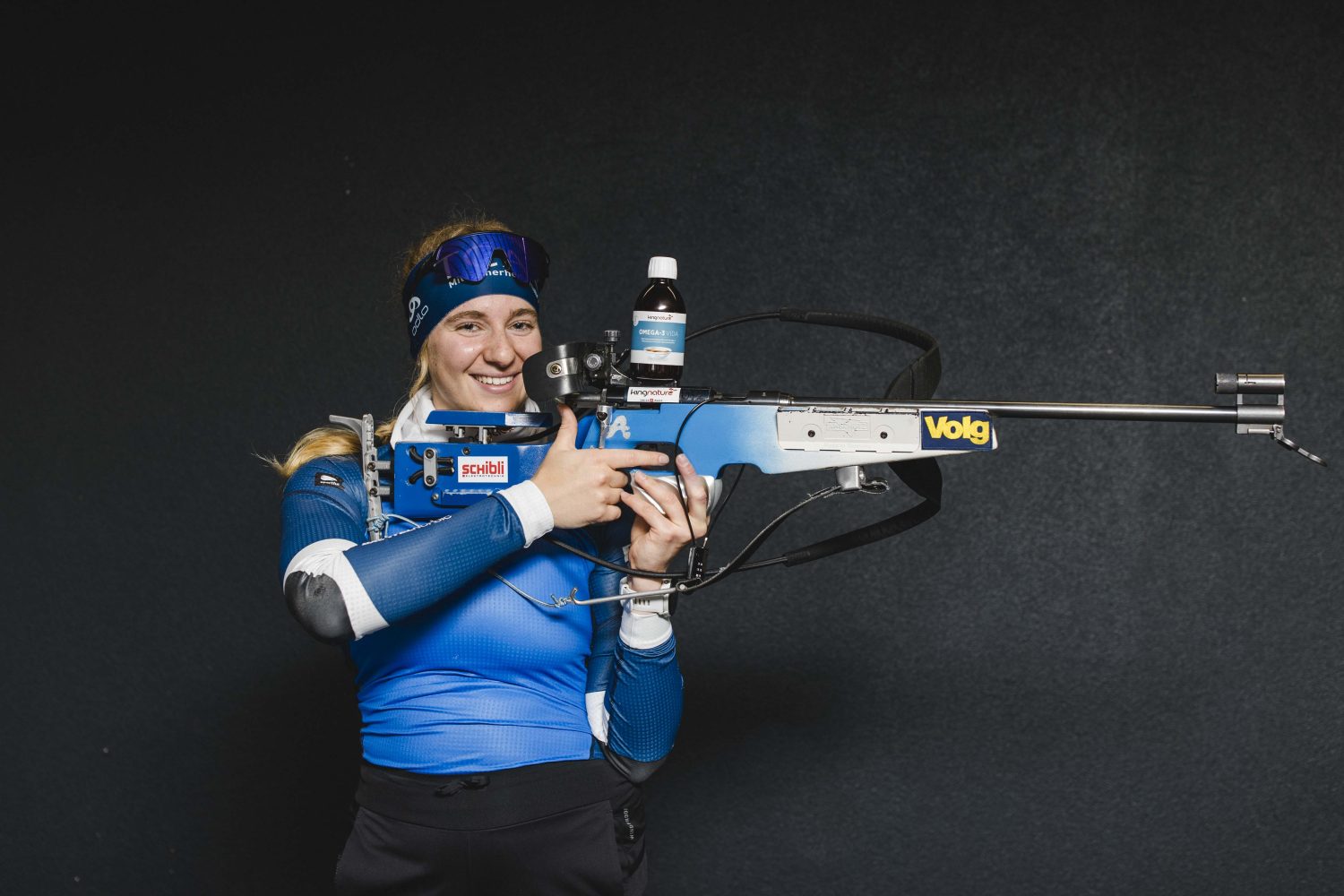 Lisa Naegeli, eine lächelnde Frau in blauem Sportoutfit und Stirnband, hält ein blau-silbernes Biathlongewehr mit Sponsorenaufklebern in der Hand und steht vor einem dunklen Hintergrund.