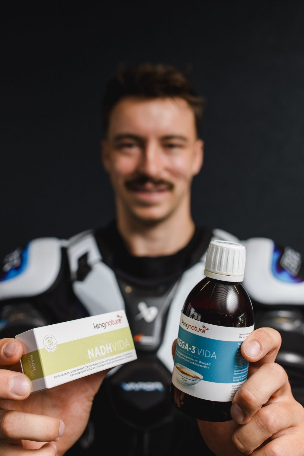Eine Person in Sportkleidung, die als Niklas Postel identifiziert wurde, hält zwei Nahrungsergänzungsmittel – NADH VIDA (Schachtel) und OMEGA-3 VIDA (Flasche) – hoch und lächelt vor einem dunklen Hintergrund in die Kamera.