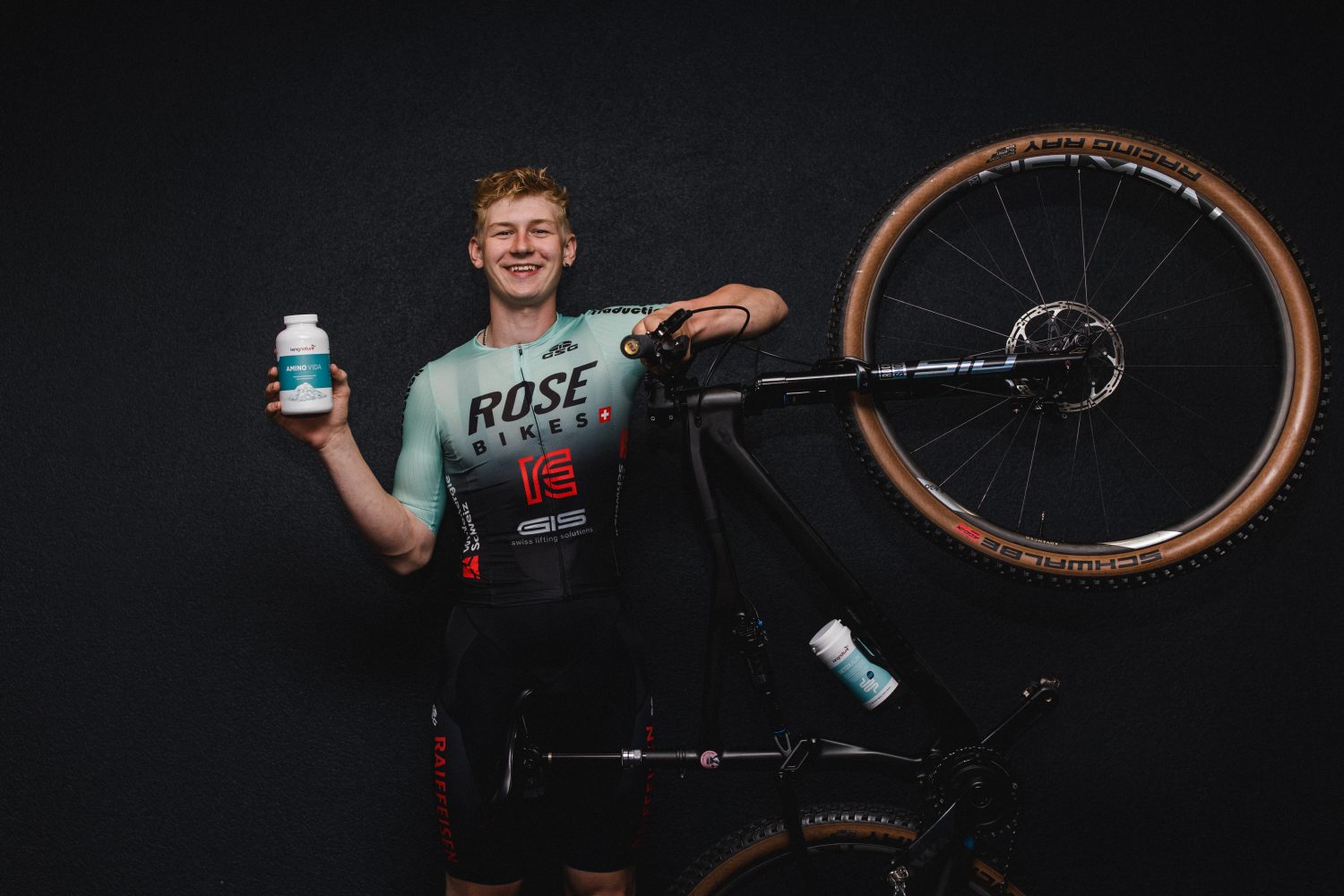 Ein Radfahrer in einem grün-schwarzen ROSE Bikes-Trikot lächelt, während er eine Trinkflasche in der Hand hält. Er steht neben einem Mountainbike, dessen eines Rad in die Höhe gereckt ist, vor einem dunklen Hintergrund – und repräsentiert stolz das Factory Racing Team.