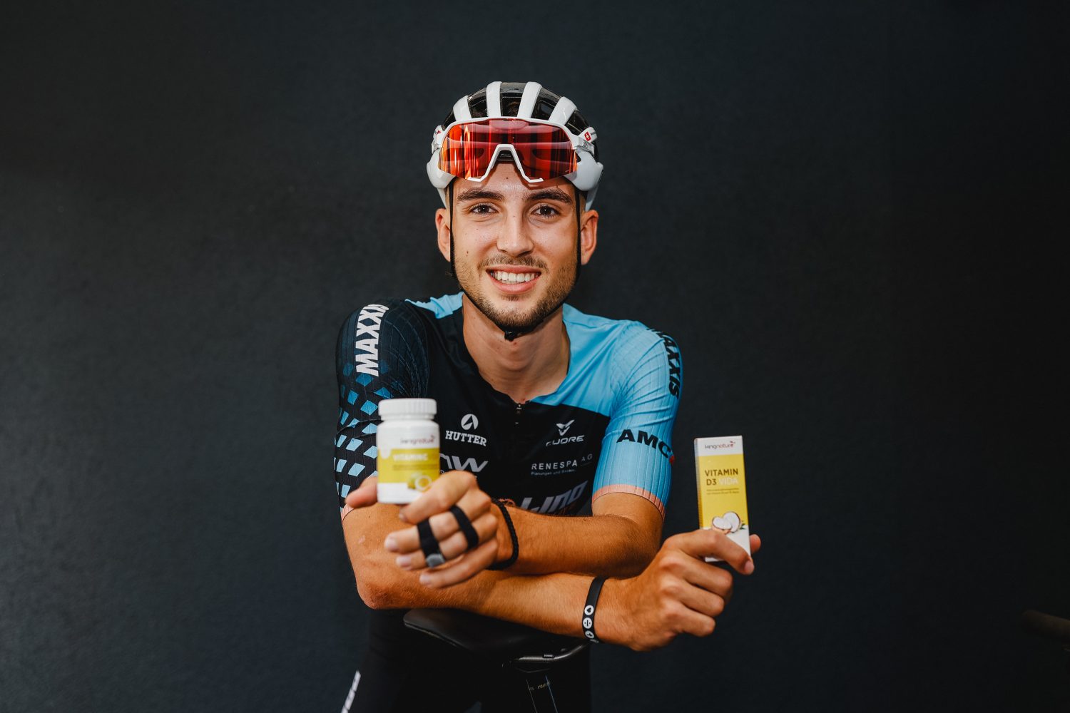 Ein lächelnder Radfahrer mit Helm und Sporttrikot lehnt sich an den Lenker seines Fahrrads. Vor einem dunklen Hintergrund hält er in der einen Hand eine Flasche Vitamin C und in der anderen eine Packung Vitamin D3 Vida.