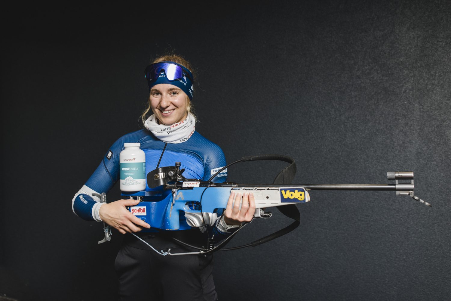 Ein lächelnder Sportler in einem blauen Sportoutfit mit Visier hält ein blaues Biathlongewehr, eine große Wasserflasche und Amino Vida-Kapseln vor einem dunklen Hintergrund.