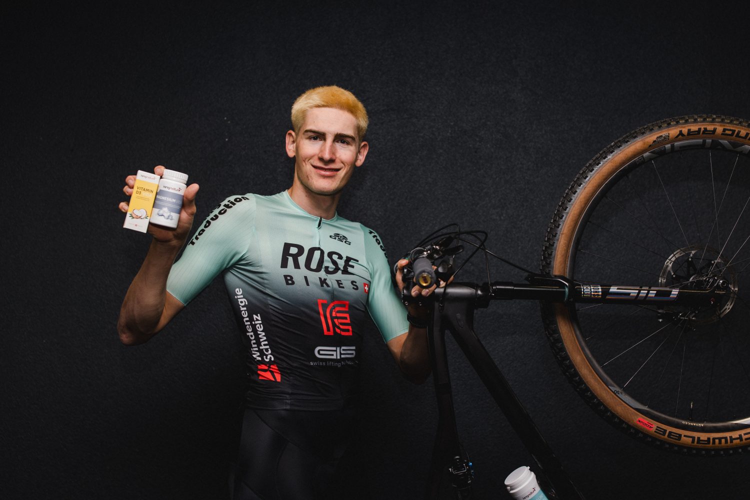 Ein Radfahrer in einem blau-schwarzen Trikot des ROSE Bikes Factory Racing Teams steht neben einem an der Wand befestigten Fahrrad, lächelt und hält zwei kleine Produktverpackungen in einer Hand hoch.