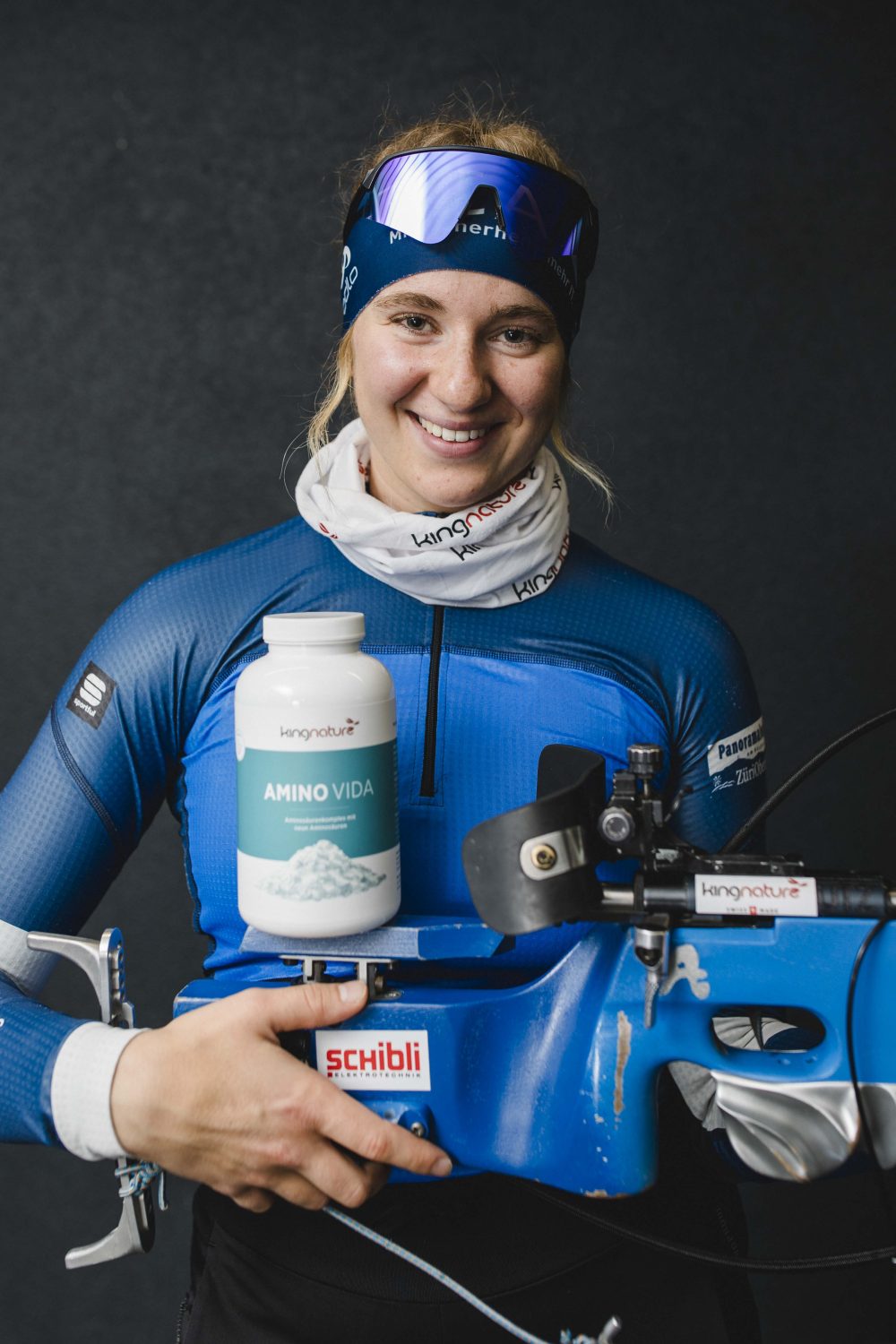 Lisa Naegeli, eine lächelnde Sportlerin in blauem Outfit, hält vor dunklem Hintergrund ein blaues Biathlongewehr und einen Behälter mit der Aufschrift „Amino Vida“. Sie trägt ein Stirnband, einen Halswärmer und Sportkleidung.