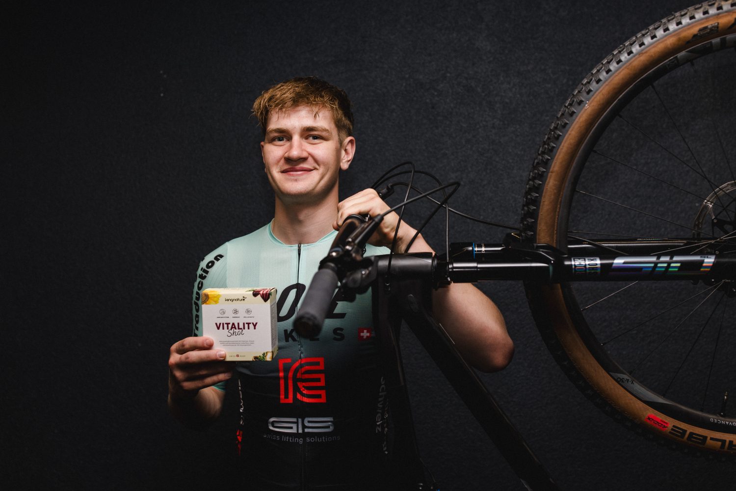 Ein lächelnder Radfahrer in der Ausrüstung des Factory Racing Teams hält eine Schachtel mit der Aufschrift „Vitality“ in der Hand, während er neben einem ROSE-Fahrrad mit einem vorne montierten Rad vor einem dunklen Hintergrund steht.