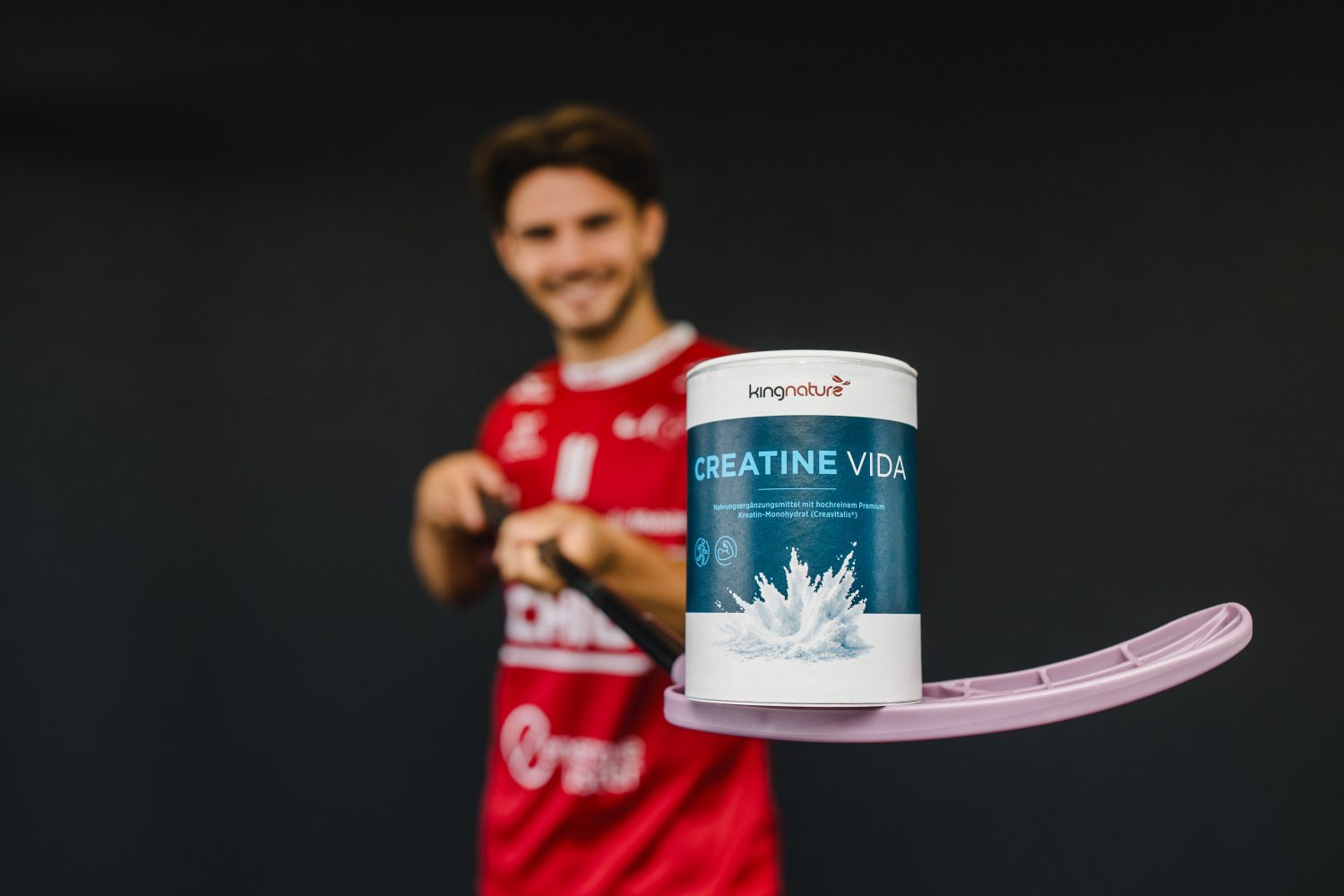 Vor einem dunklen Hintergrund hält ein Mann in einem roten Sporttrikot, der als Josia Pfister identifiziert wurde, einen Hockeyschläger in der Hand, auf dessen Klinge ein Behälter mit dem Nahrungsergänzungsmittel „Creatine Vida“ balanciert.