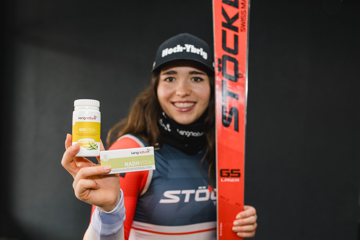 Eine Skifahrerin in Rennkleidung lächelt in die Kamera, in der Hand eine Flasche NADH Vida-Präparate und einen roten Stöckli-Ski.