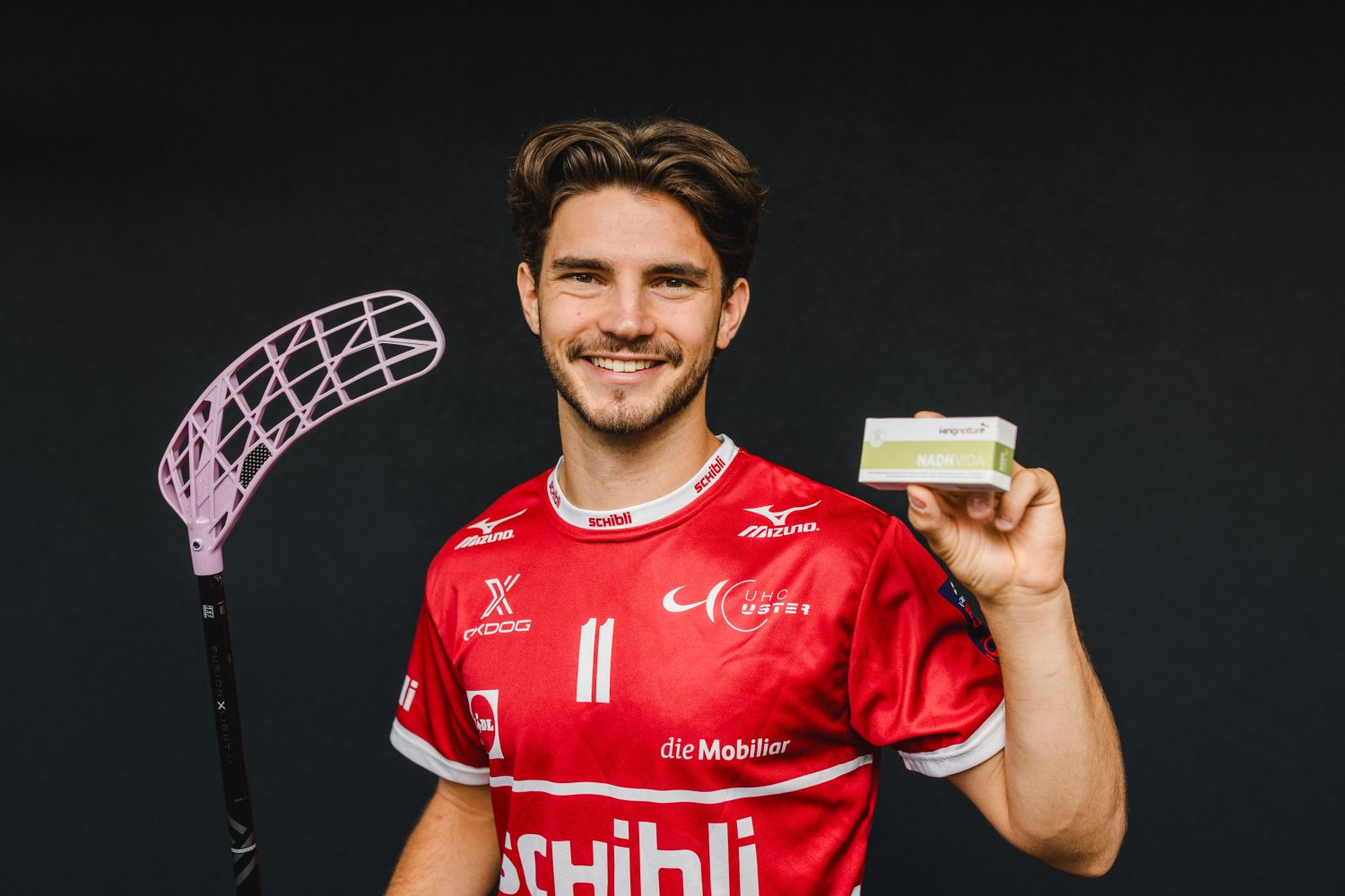Ein lächelnder Mann in einem roten Sporttrikot, der als Josia Pfister identifiziert wurde, hält in der einen Hand einen Floorballschläger und in der anderen eine kleine Schachtel und steht vor einem dunklen Hintergrund.