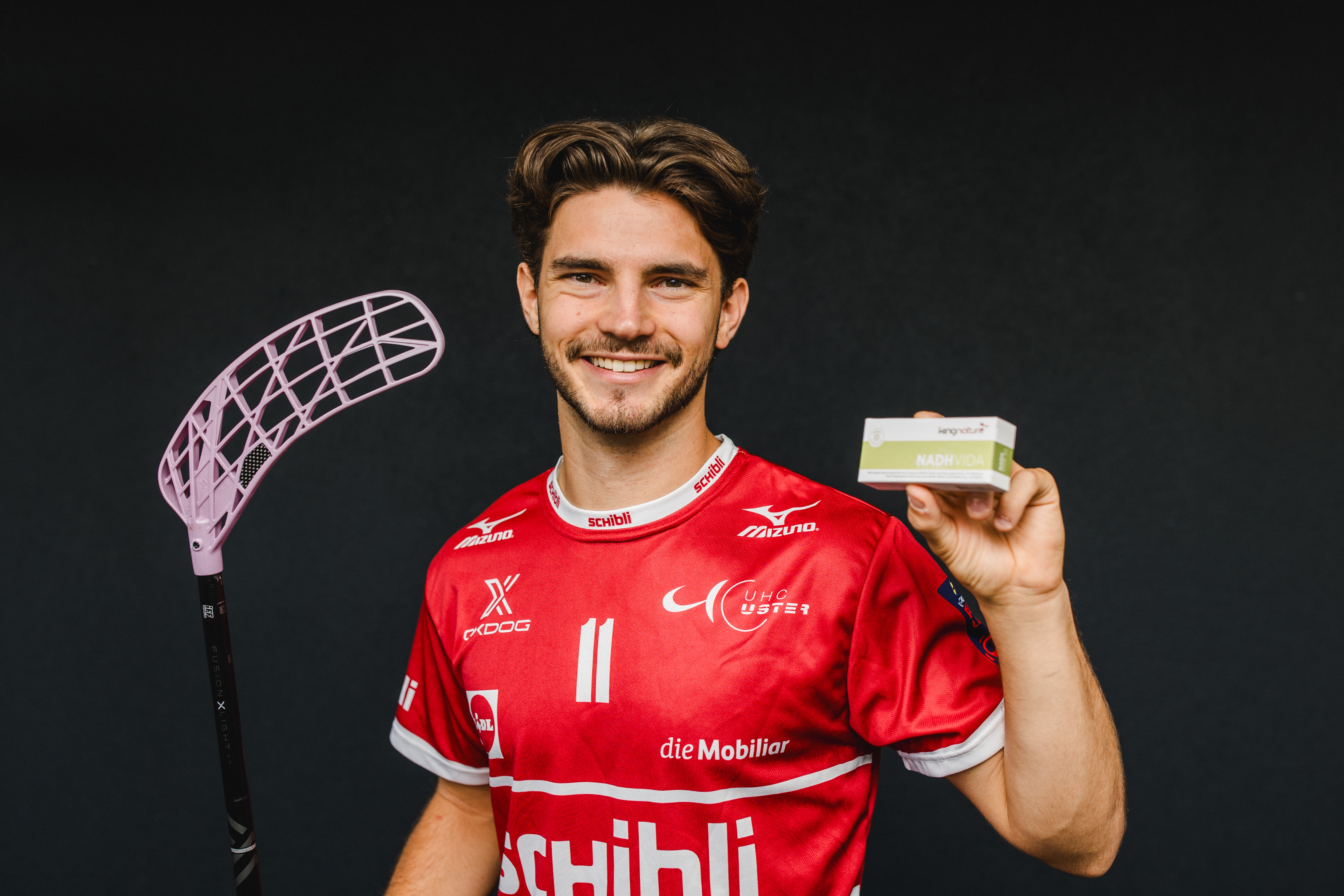 Ein lächelnder Mann in einem roten Sporttrikot, der als Josia Pfister identifiziert wurde, hält in der einen Hand einen Floorballschläger und in der anderen eine kleine Schachtel und steht vor einem dunklen Hintergrund.