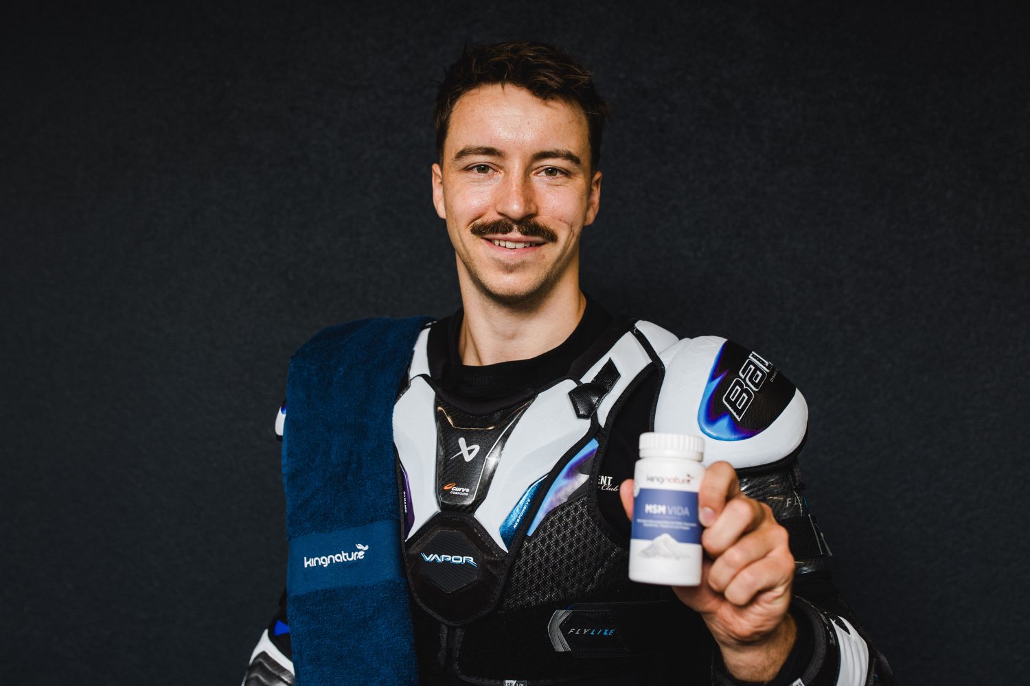 Ein Mann in Eishockeyausrüstung, der ein blaues Handtuch über die Schulter geworfen hat, lächelt und hält vor einem dunklen Hintergrund eine Flasche mit dem Nahrungsergänzungsmittel „High Vital“ in die Kamera – es ist Niklas Postel, der sein Lieblingsprodukt präsentiert.