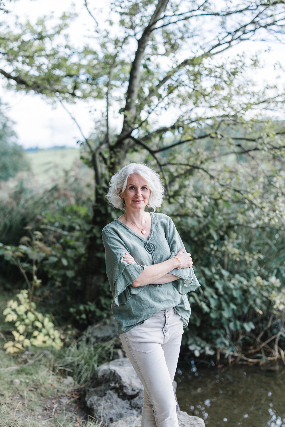 Eine Frau mit lockigen grauen Haaren, in einem grünen Oberteil und einer hellen Hose, steht mit verschränkten Armen an einem belaubten Baum in der Nähe eines Gewässers - im Freien an einem bewölkten Tag - und ruft den Geist der Gesunden Medizin für Zeiten wie diese hervor.