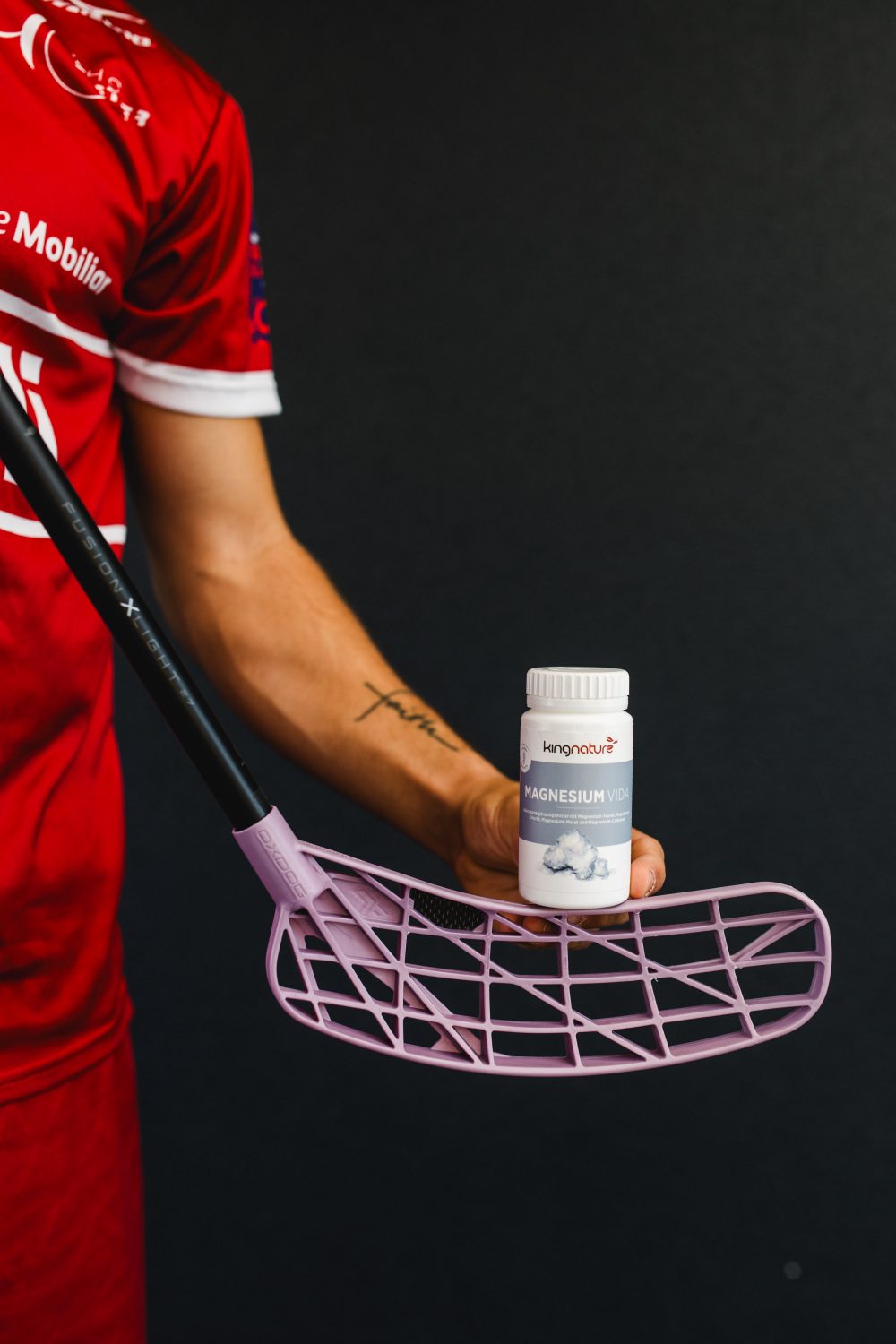 Vor einem dunklen Hintergrund hält eine Person in einem roten Sportoutfit einen Floorballschläger mit hellviolettem Blatt in der Hand und balanciert darauf eine Flasche mit der Aufschrift „Magnesium“ – ein Bild, das das Können und die Konzentration von Josia Pfister perfekt einfängt.
