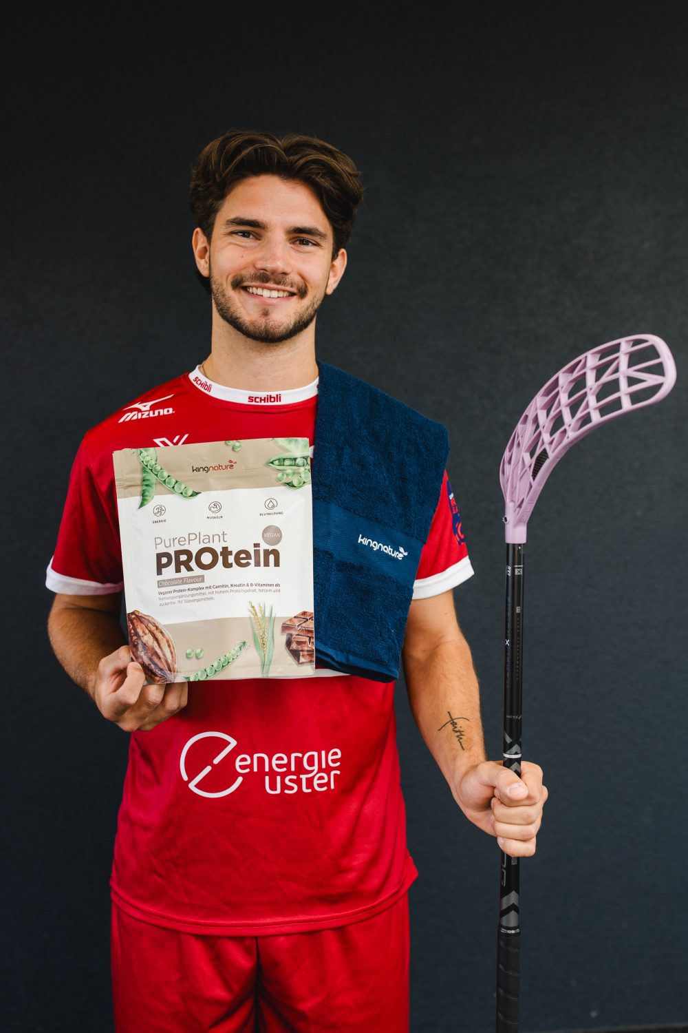 Ein lächelnder Sportler in einem roten Trikot, der an Josi Pfister erinnert, hält einen Floorballschläger und eine Tüte Proteinpulver in der Hand, hat ein dunkelblaues Handtuch über die Schulter geworfen und steht vor einem dunklen Hintergrund.