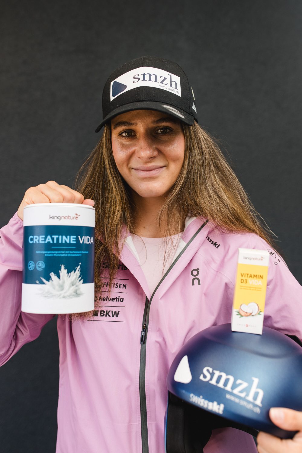 Talina Gantenbein, die eine rosa Jacke und eine schwarze Mütze trägt, lächelt, während sie einen Behälter mit der Aufschrift „CREATINE VIDA“ und einen blauen Helm mit einer Schachtel mit der Aufschrift „VITAMIN D3 5000IU“ in die Höhe hält; sie steht vor einem dunklen Hintergrund.