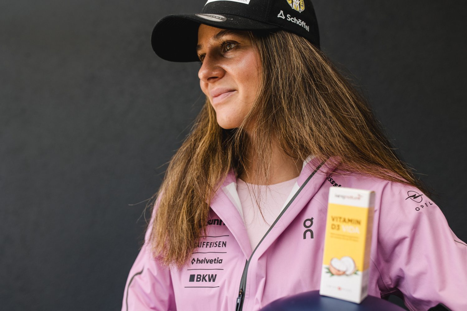 Eine lächelnde Frau in einer rosa Sportjacke und einer schwarzen Mütze steht vor einem dunklen Hintergrund. Vor ihr steht eine Schachtel mit der Aufschrift „Vitamin D3 Vida“, die Talina Gantenbeins Entscheidung für einen gesunden Lebensstil unterstreicht.