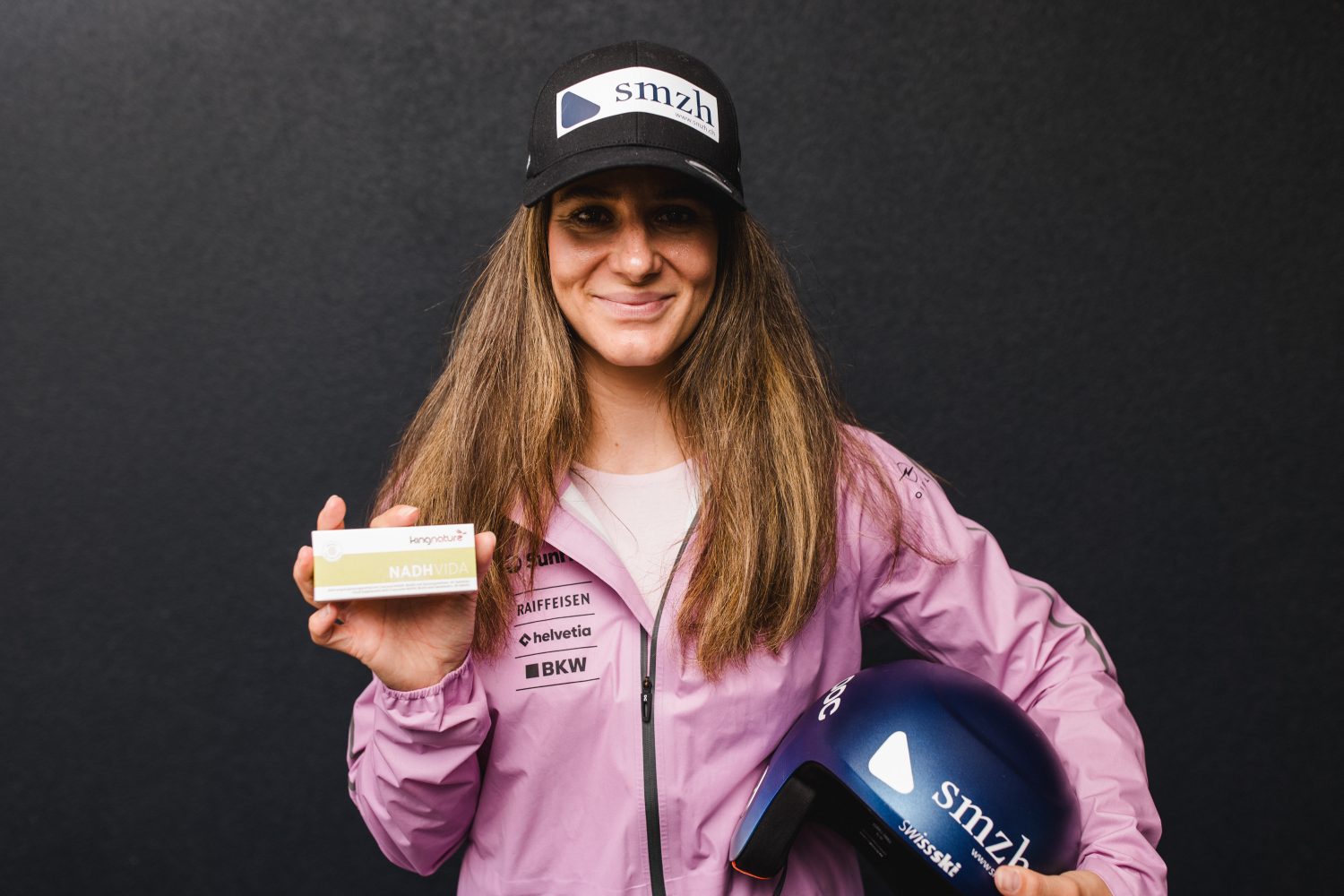Eine Frau, die als Talina Gantenbein identifiziert wurde, trägt eine rosa Jacke und eine schwarze Mütze mit dem „smzh“-Logo. Sie lächelt, während sie in der einen Hand einen blauen Helm und in der anderen eine Schachtel mit der Aufschrift „NADH 100“ hält, und steht vor einem schlichten dunklen Hintergrund.