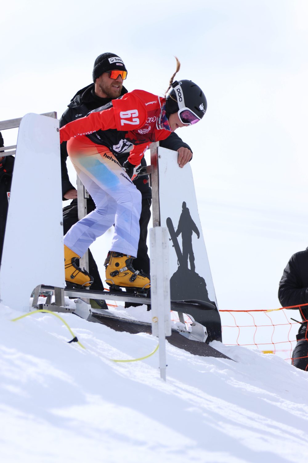 Eine Snowboarderin in weißer Hose und rotem Trikot, mit Helm und Schutzbrille, bereitet sich auf den Start eines Rennens vor. Ein Offizieller assistiert ihr am verschneiten Starttor – Ricarda Hauser steht daneben. Ein orangefarbenes Netz markiert die Strecke.