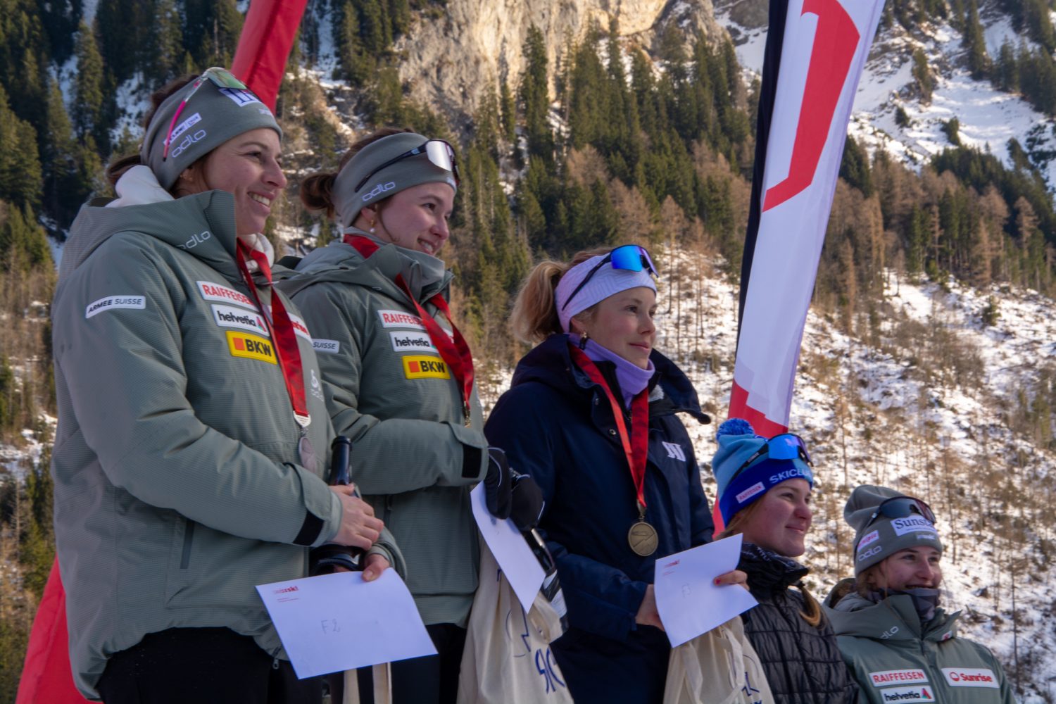 Fünf Skifahrer in Winterkleidung, darunter Ramona Schöpfer, stehen mit Medaillen und Umschlägen auf einem Podium im Freien und lächeln. Im Hintergrund sind schneebedeckte Bäume und Berge zu sehen, daneben Fahnen. Es scheint sich um eine Siegerehrung eines Skiwettbewerbs zu handeln.
