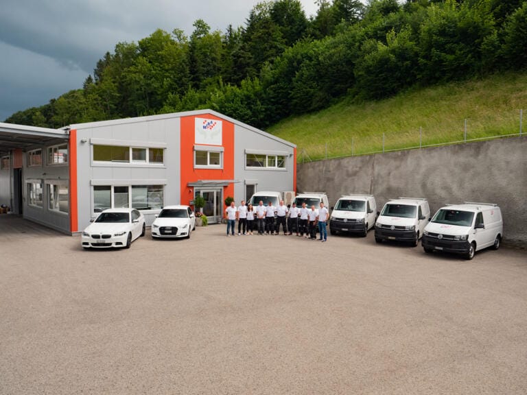 Eine Gruppe von Menschen in weißen Hemden steht vor einem modernen Gebäude mit orangefarbenen Akzenten. Mehrere weiße Autos und Lieferwagen parken in der Nähe, neben Grünflächen und einem Hügel, was eine organisierte Teamatmosphäre widerspiegelt.