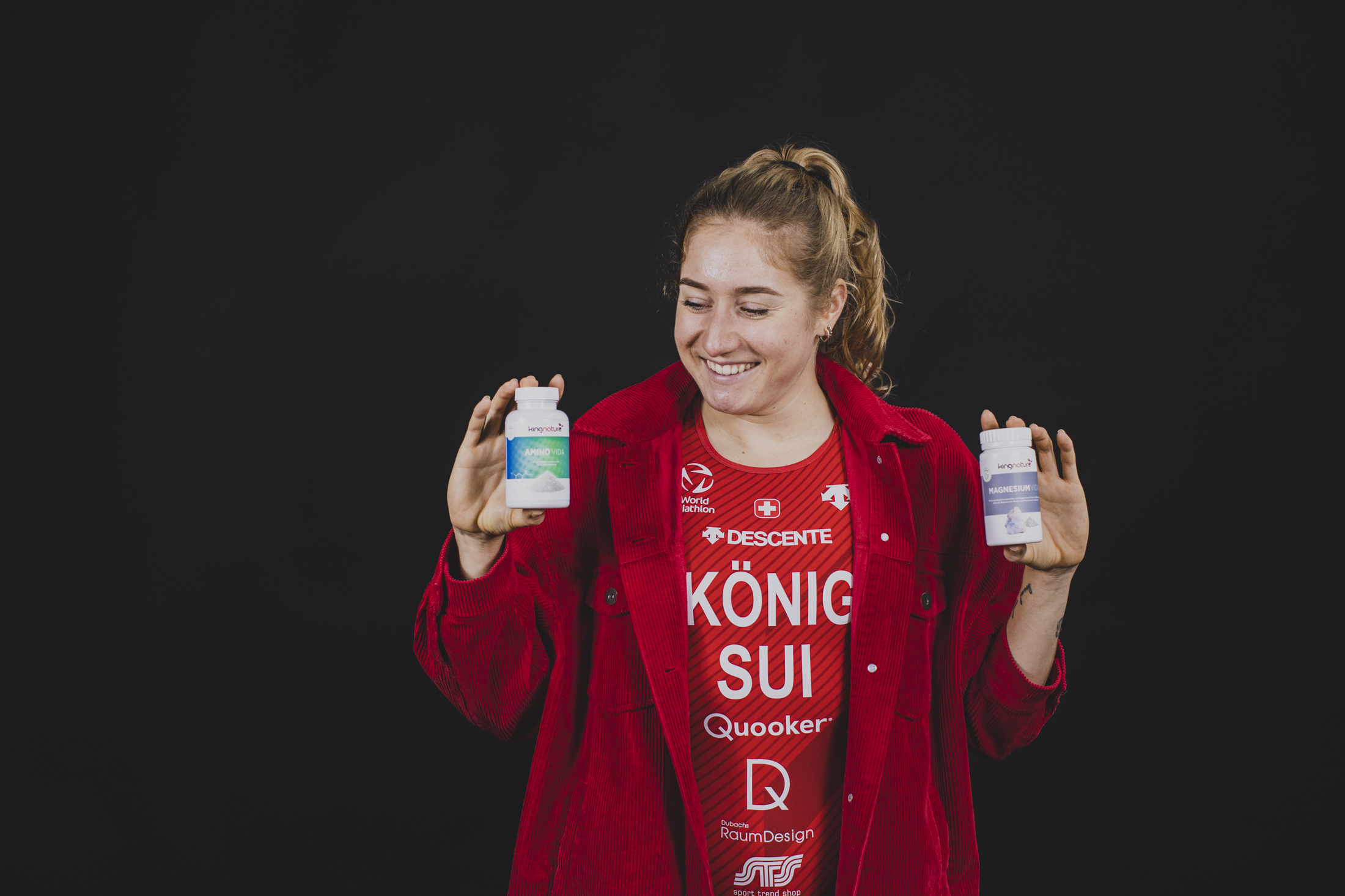 A smiling woman in athletic wear holds a tennis racket and a bottle of supplements against a dark background.