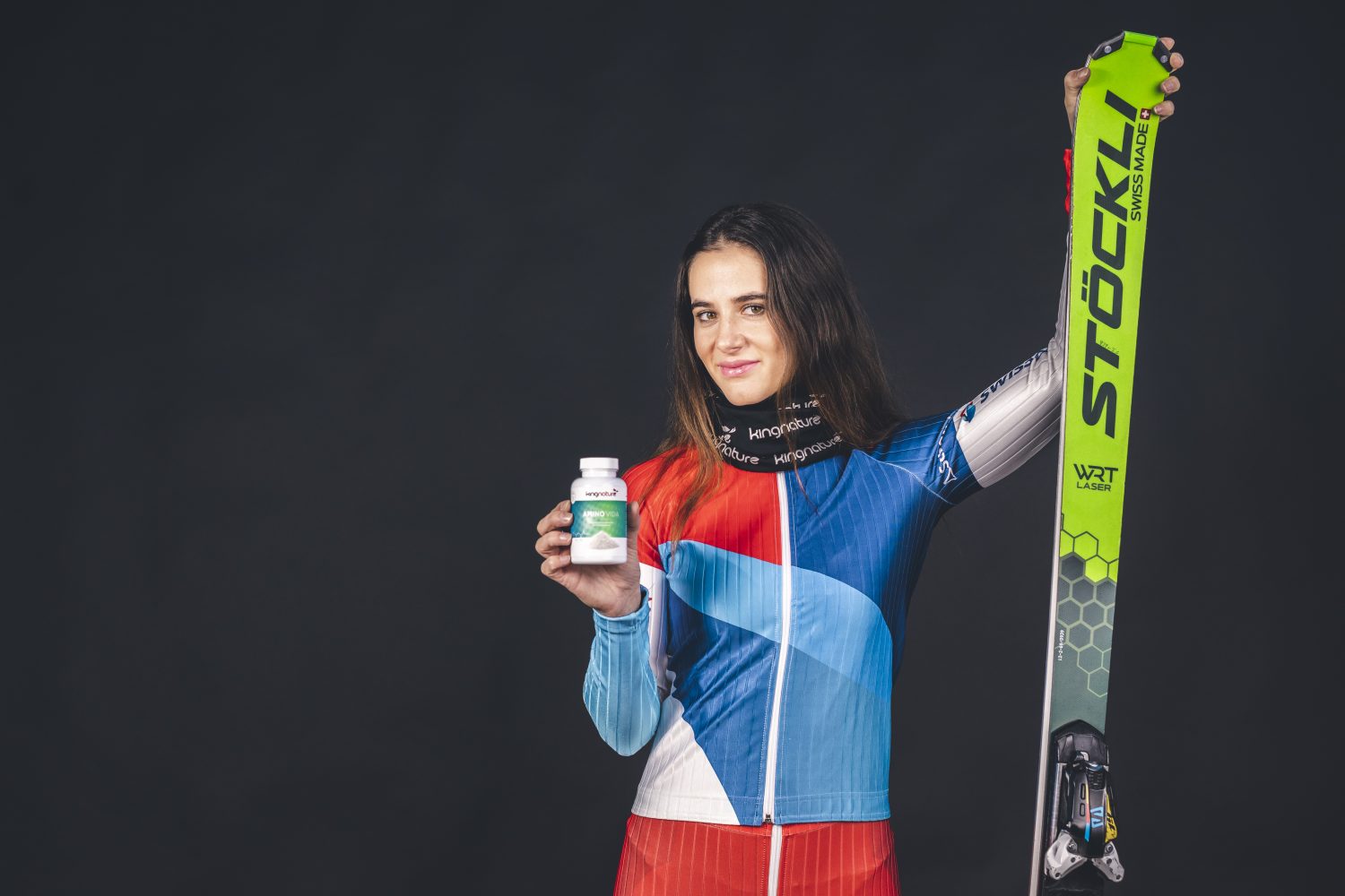 Eine Skifahrerin im bunten Rennanzug steht vor einem dunklen Hintergrund und hält in der einen Hand einen grünen Stöckli-Ski und in der anderen eine Flasche Amino Vida-Präparate.