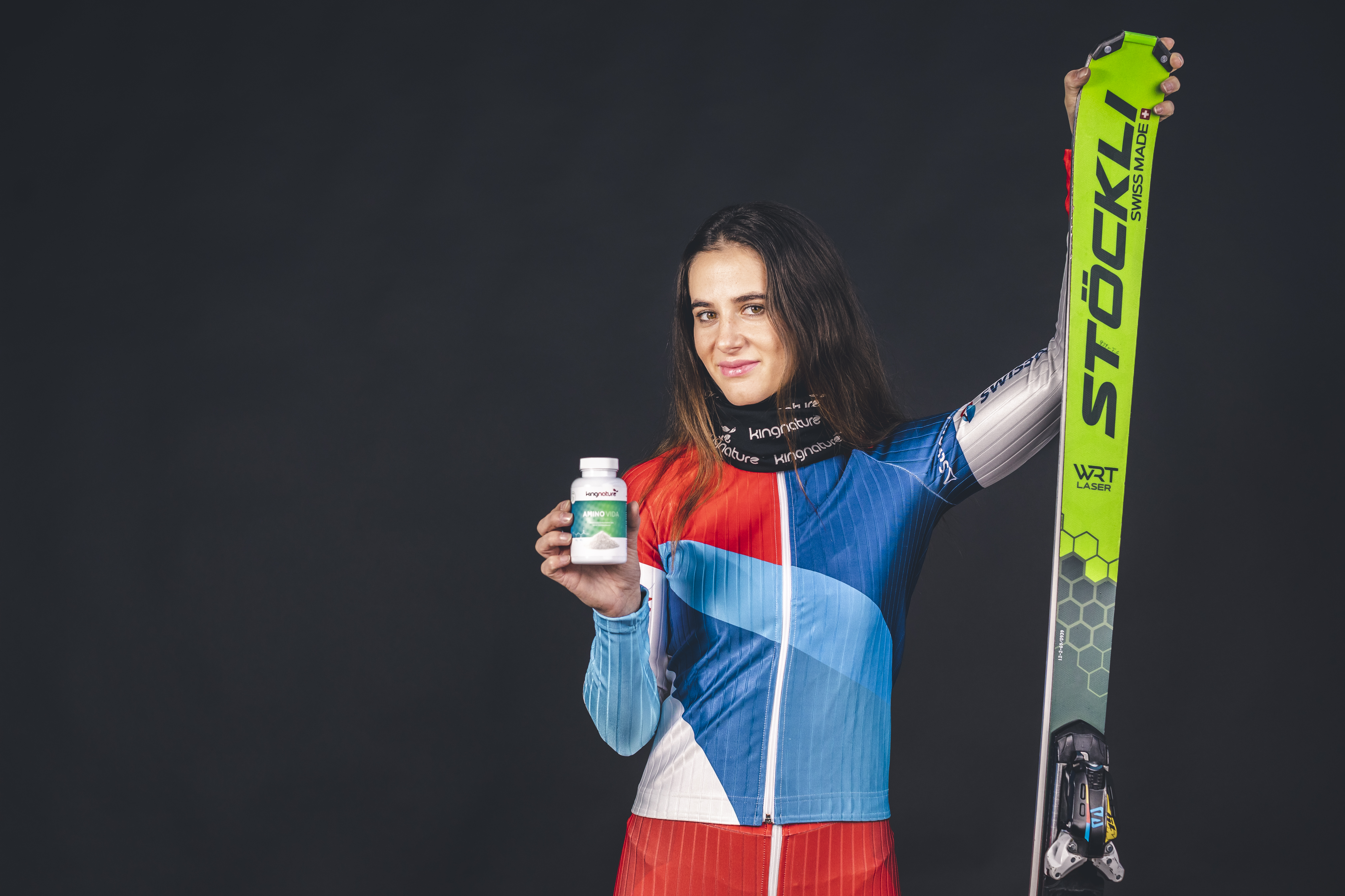 Eine Skifahrerin im bunten Rennanzug steht vor einem dunklen Hintergrund und hält in der einen Hand einen grünen Stöckli-Ski und in der anderen eine Flasche Amino Vida-Präparate.