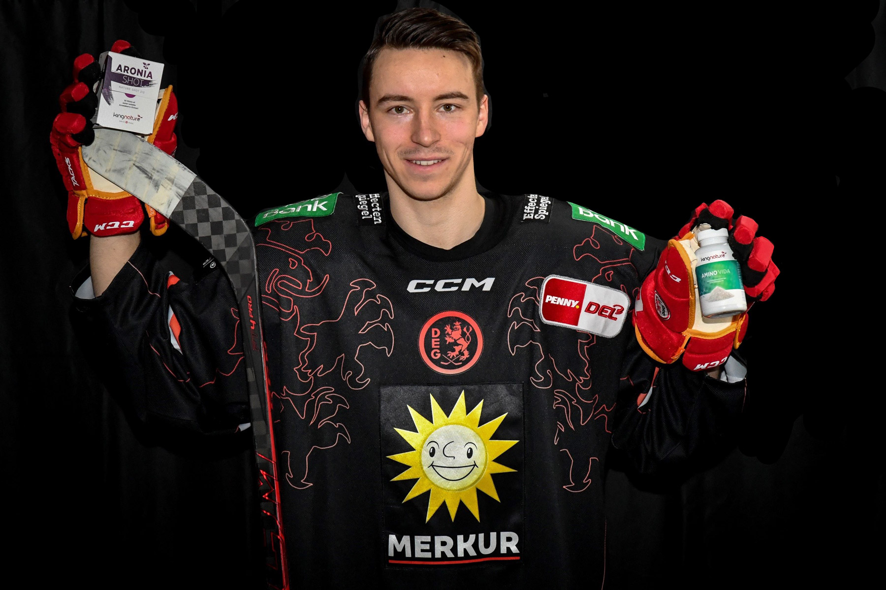 Ein Hockeyspieler in einem schwarzen Trikot mit Sonnenlogo und Sponsorenlogos, der rote Handschuhe trägt, lächelt in die Kamera, während er in der einen Hand einen Hockeyschläger und in der anderen zwei Amino Vida-Flaschen hält.