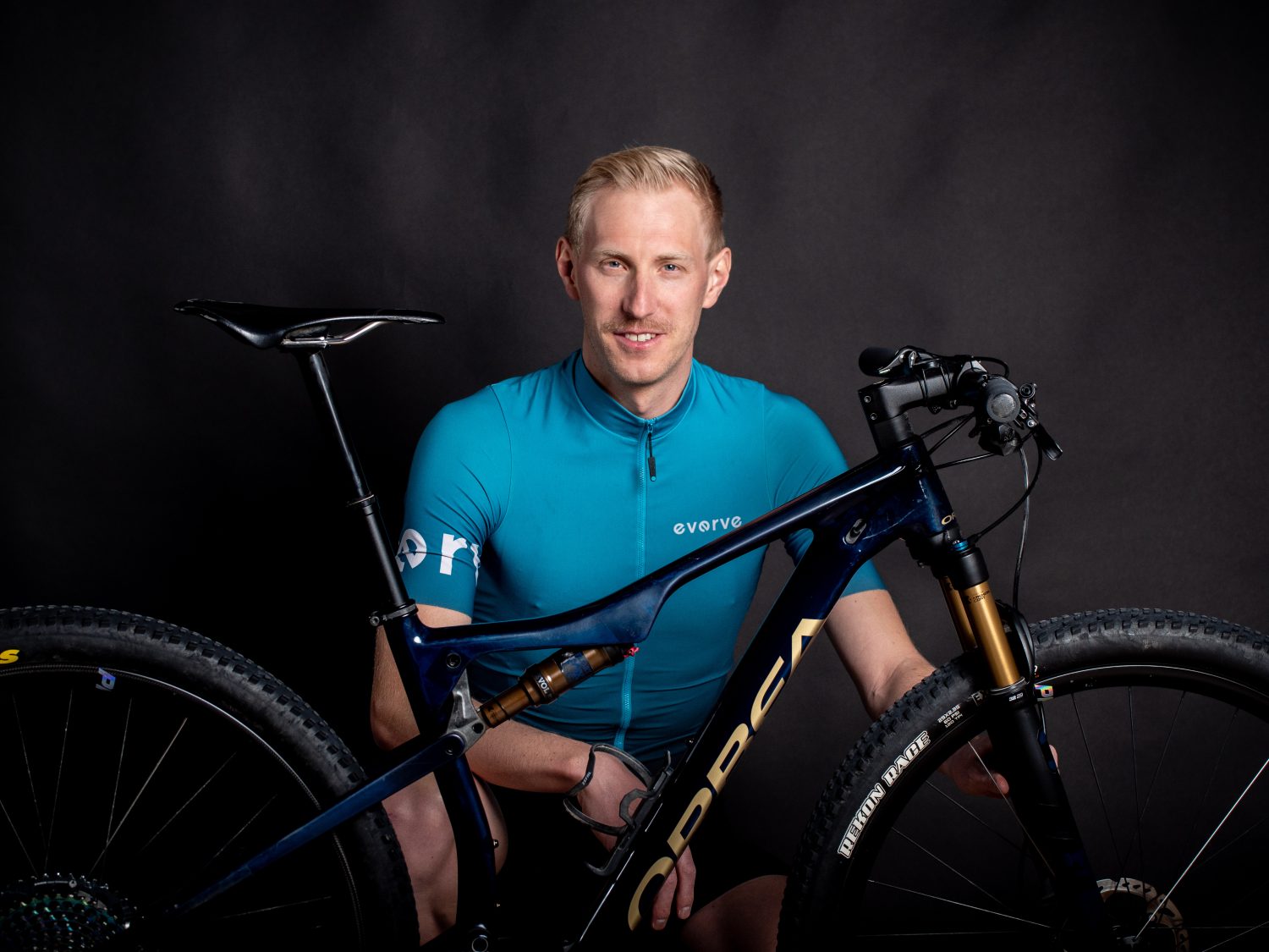 Ein Mann in einem blaugrünen Radtrikot von Amino Vida posiert mit einem dunkelblauen Orbea Mountainbike vor einem schwarzen Hintergrund und lächelt in die Kamera.
