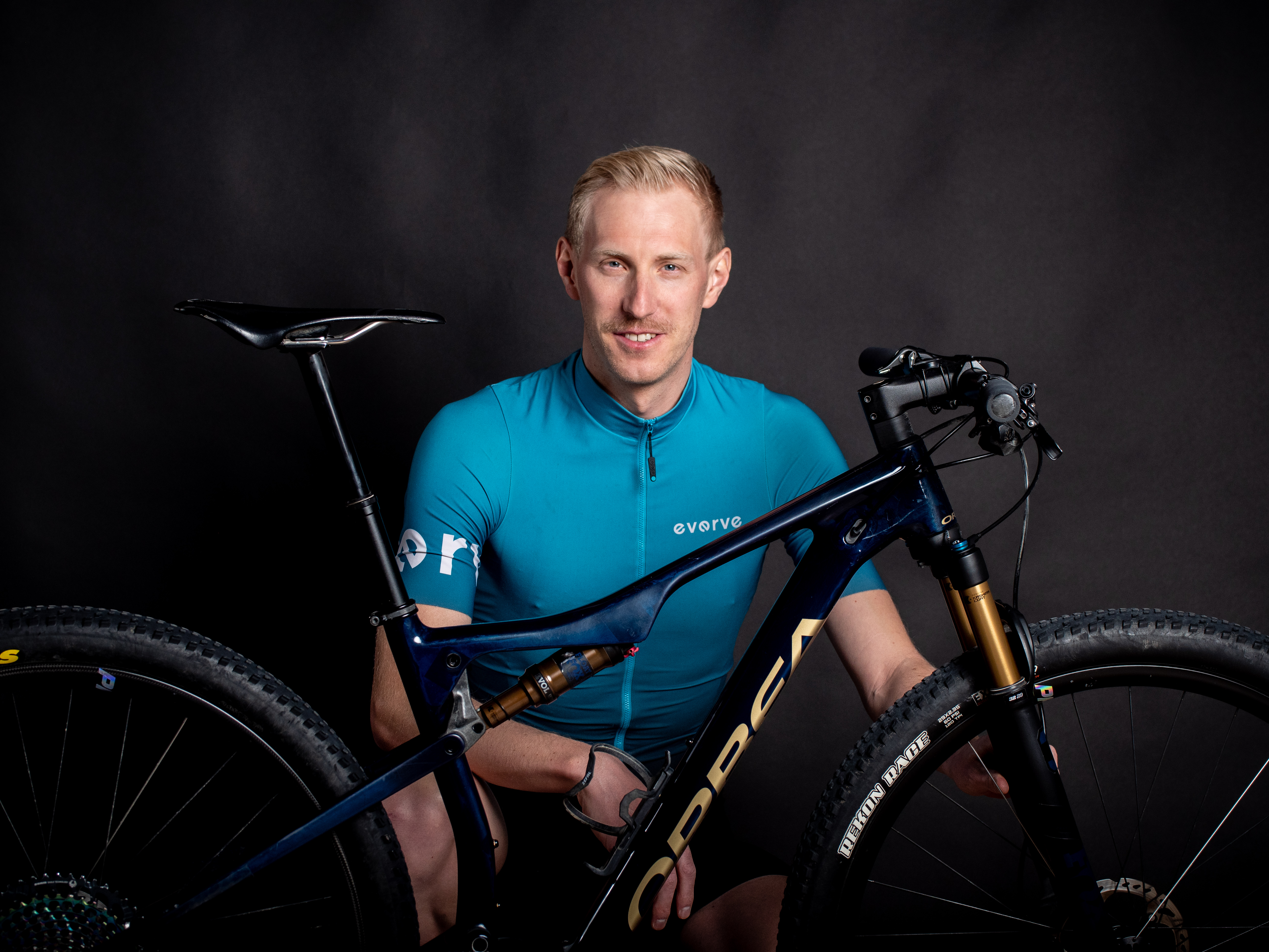 Ein Mann in einem blaugrünen Radtrikot von Amino Vida posiert mit einem dunkelblauen Orbea Mountainbike vor einem schwarzen Hintergrund und lächelt in die Kamera.