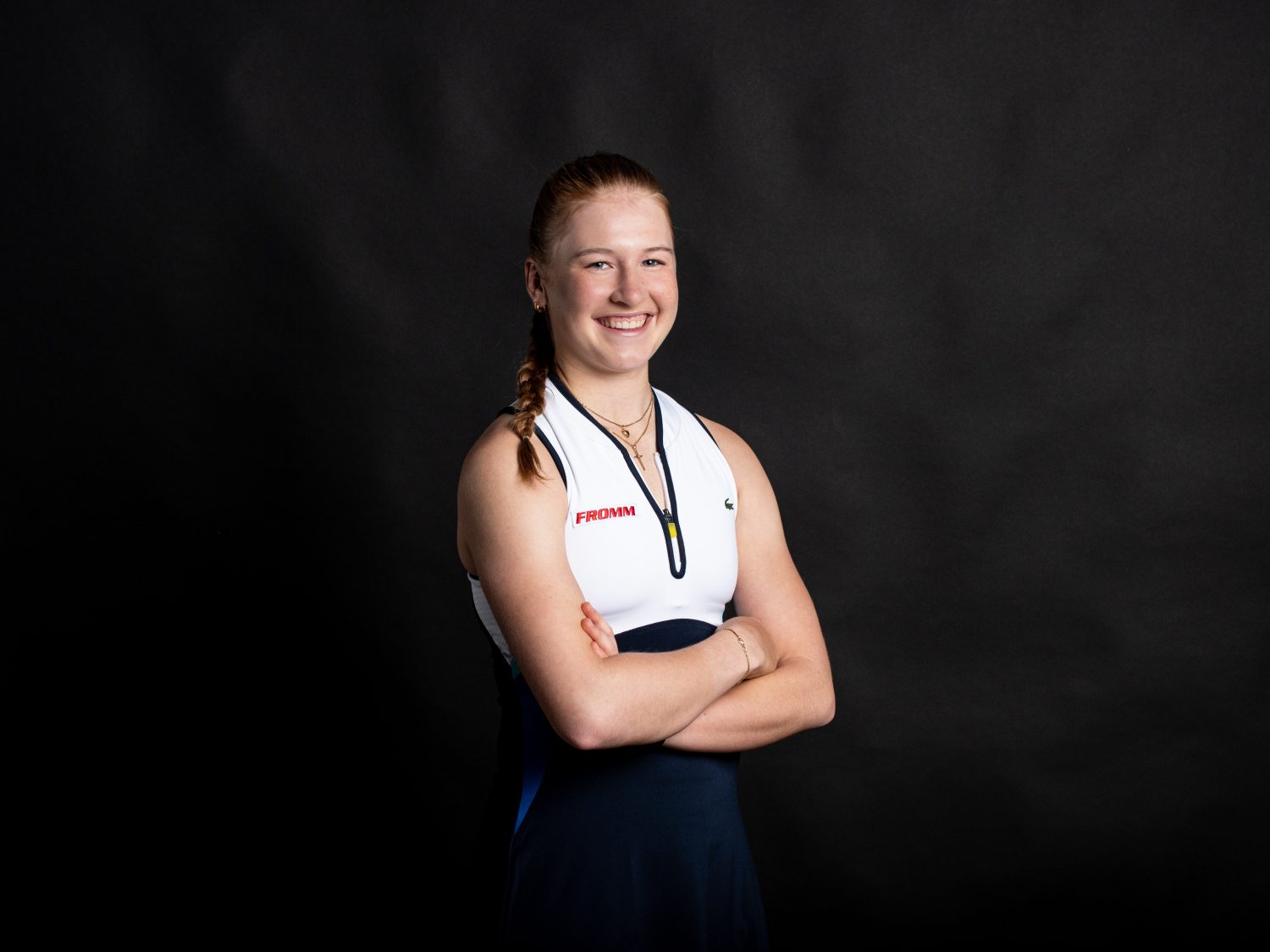 A young woman with a braid smiles confidently with her arms crossed, wearing a sleeveless athletic uniform against a dark, plain background.