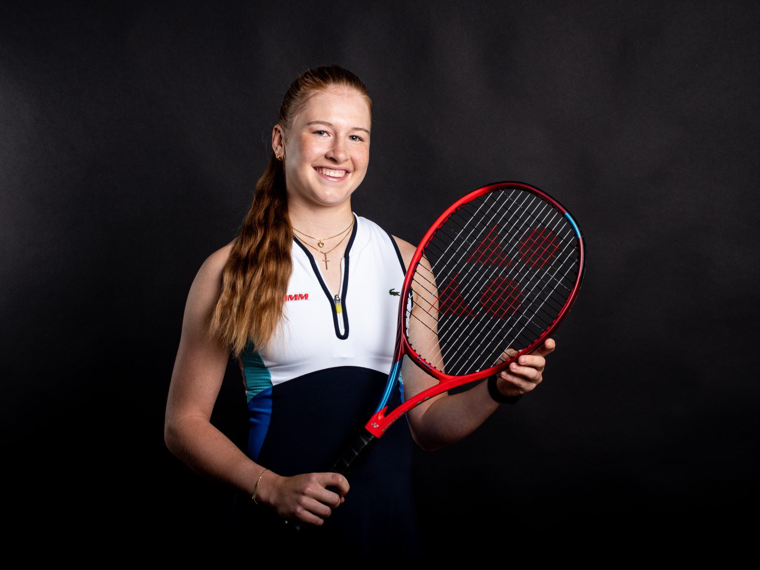 A young woman with long red hair in a ponytail, wearing a navy and white tennis outfit, smiles and holds a red and black tennis racket in front of a dark background.