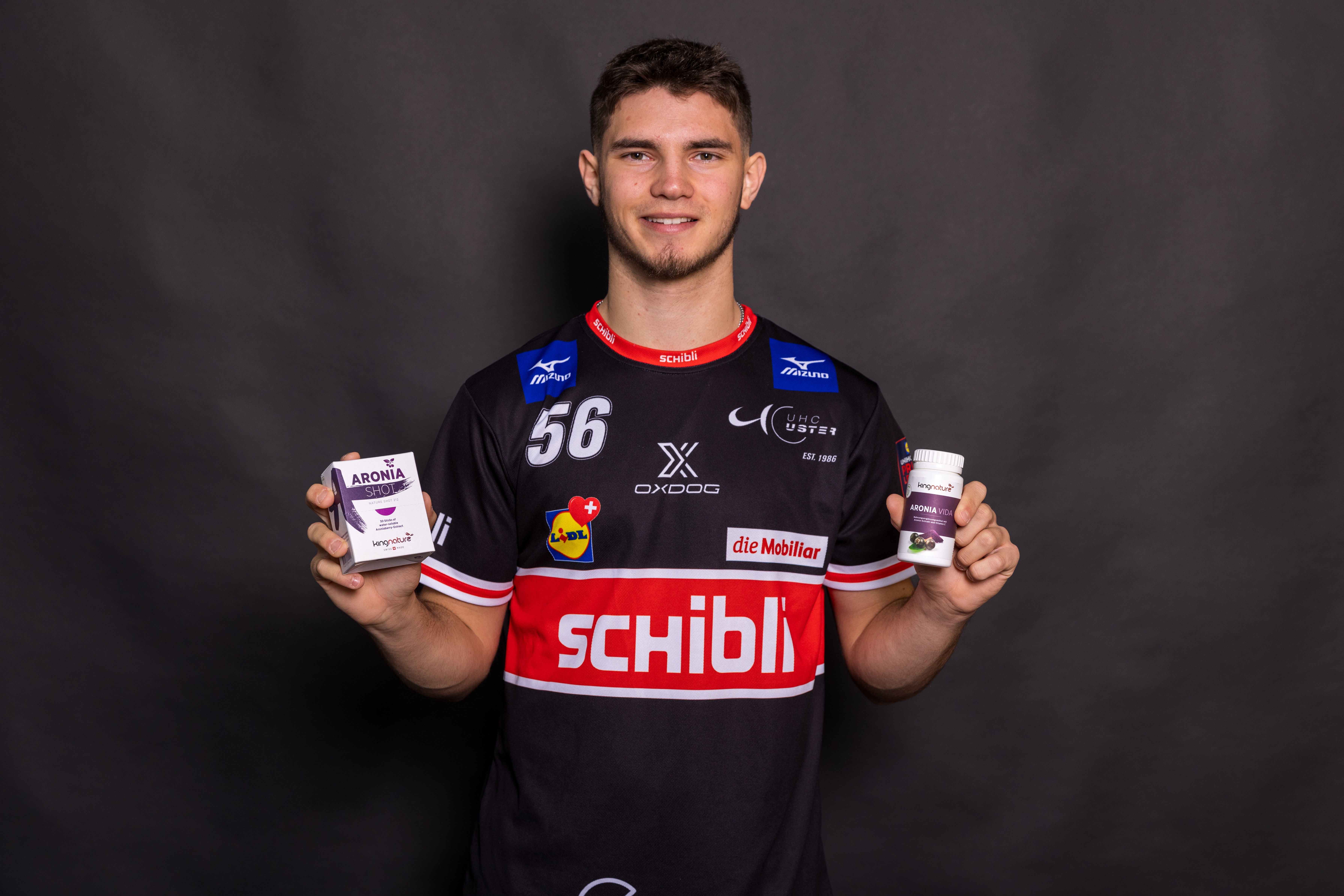 Ein junger Mann in einem schwarzen Sporttrikot mit roten und weißen Akzenten lächelt, während er zwei Flaschen Aronia Vida - ideal für alle, die Aronia Kapseln kaufen möchten - vor einem schlichten dunklen Hintergrund hält.