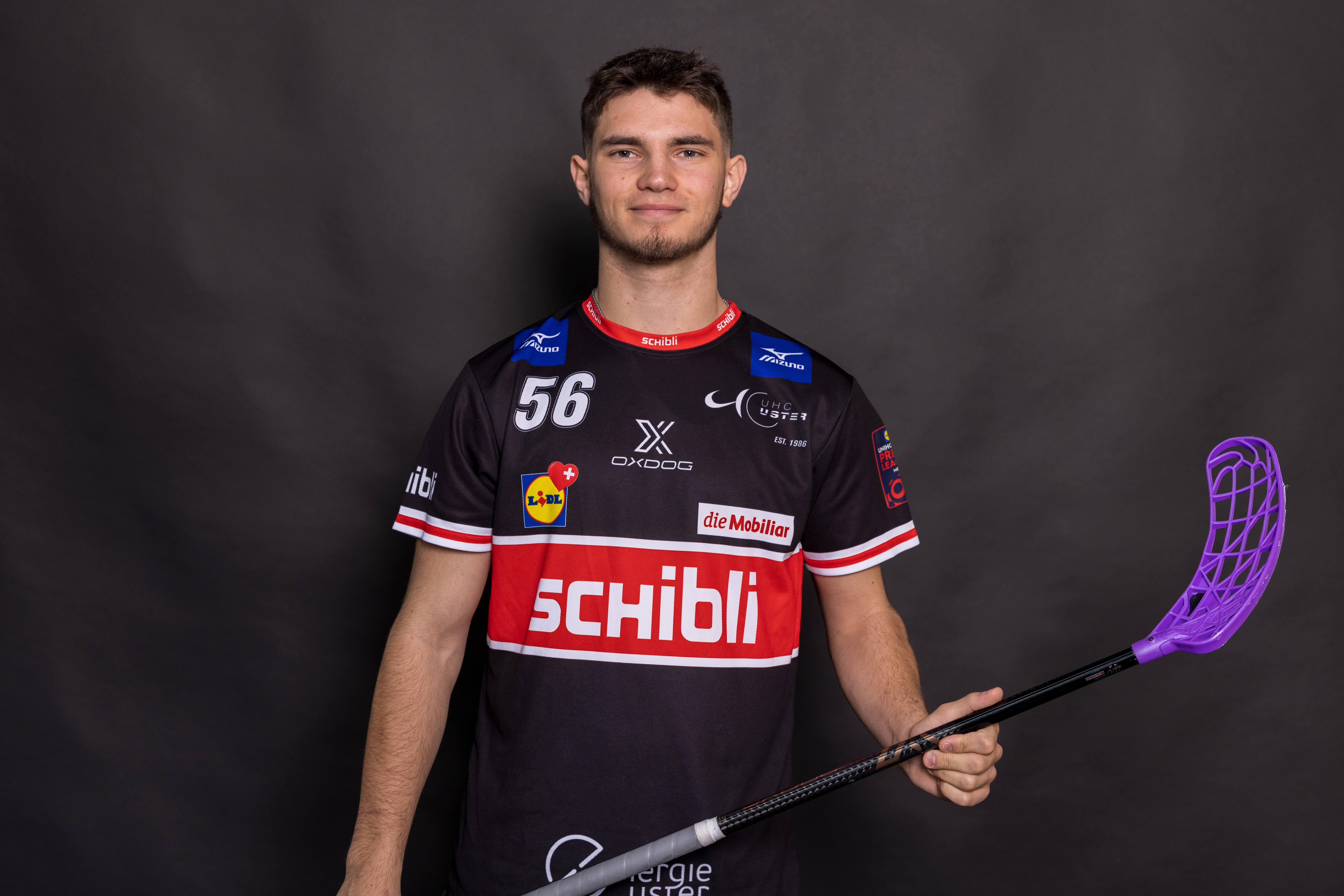 Ein junger Mann in einem schwarzen Sporttrikot mit roten und weißen Akzenten hält einen lila Unihockeyschläger und eine Flasche Amino Vida in der Hand. Er steht vor einem schlichten dunklen Hintergrund und lächelt in die Kamera.