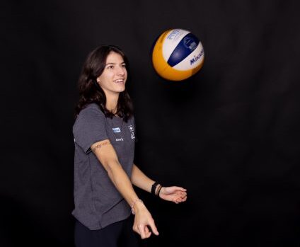 Eine Frau in grauem T-Shirt und schwarzer Hose lässt vor schwarzem Hintergrund einen Volleyball auf ihrem Finger kreisen. Sie lächelt und ist voller Energie - vielleicht hat sie sich für Moringa Vida Bio entschieden, um einen gesunden Kick zu bekommen.