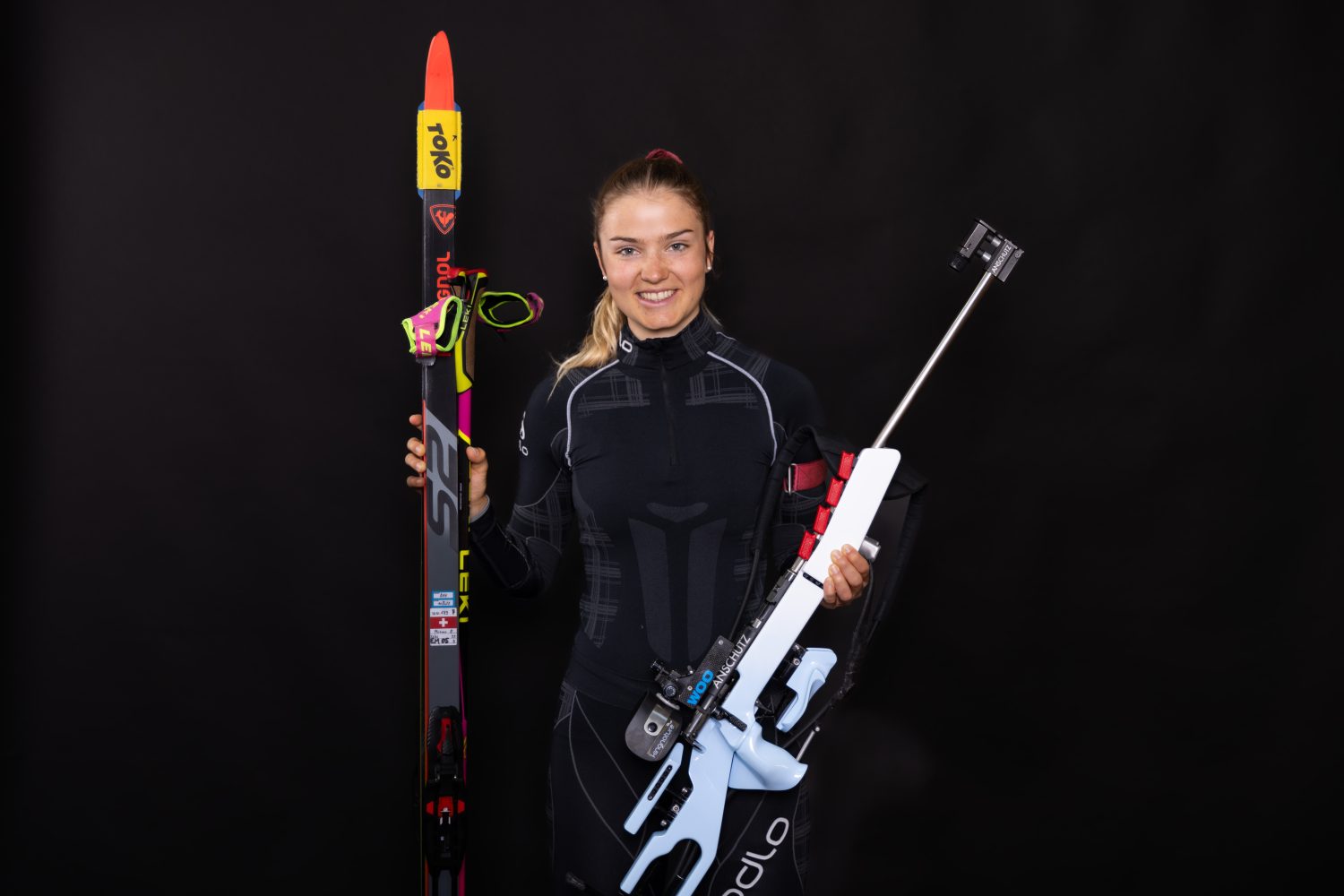 Eine Frau in einem schwarzen Sportanzug steht vor einem schwarzen Hintergrund und lächelt. Sie hält Skier und ein Biathlon-Gewehr in der Hand und verkörpert Stärke - genau wie diejenigen, die Artemisia PET Bio für natürliches Wohlbefinden wählen.